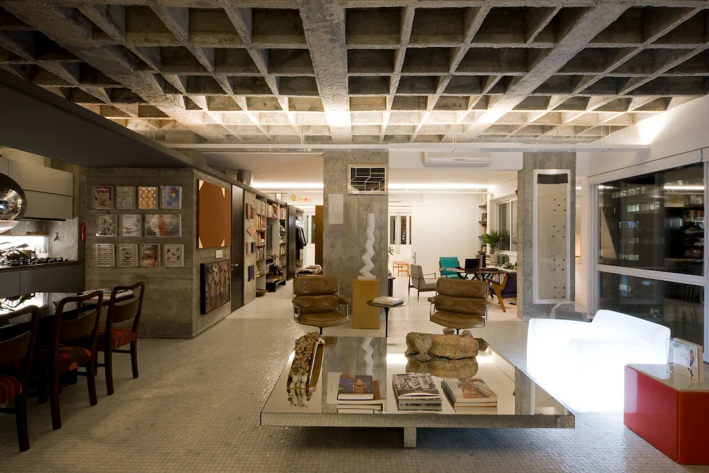 An industrial living room features an exposed concrete waffle ceiling, a large mirrored coffee table, and mid-century leather lounge chairs.