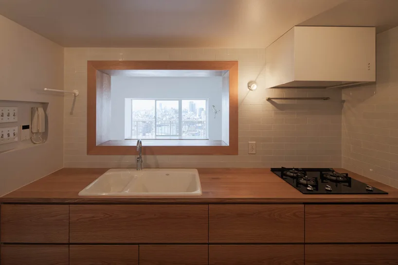 A minimalist kitchen features wood countertops and cabinetry, a white subway tile backsplash, and a rectangular window cutout showing a city view.