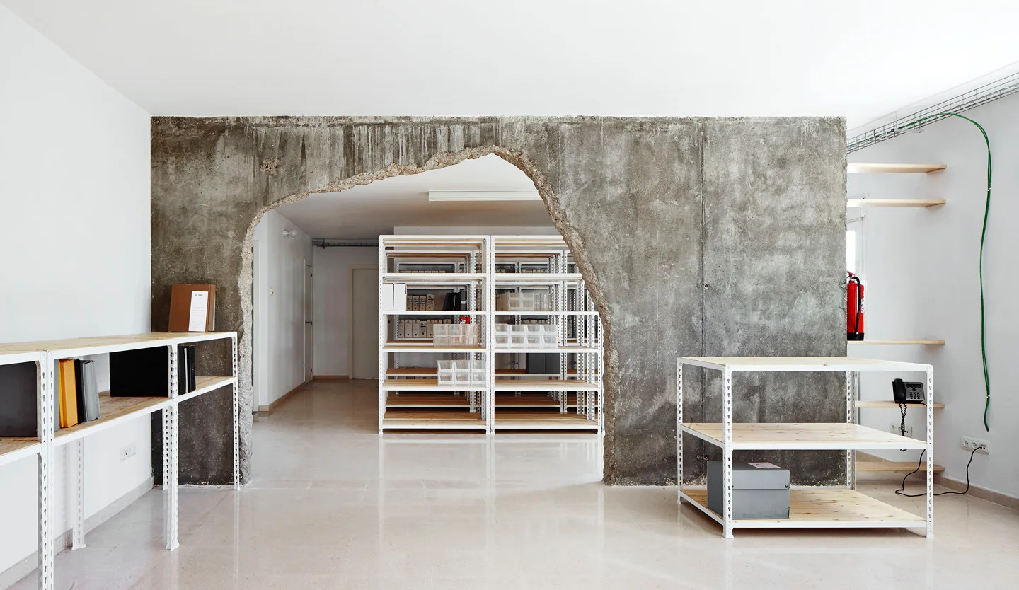 A central raw concrete partition wall with a rough arched cutout stands between minimalist white metal shelving units.