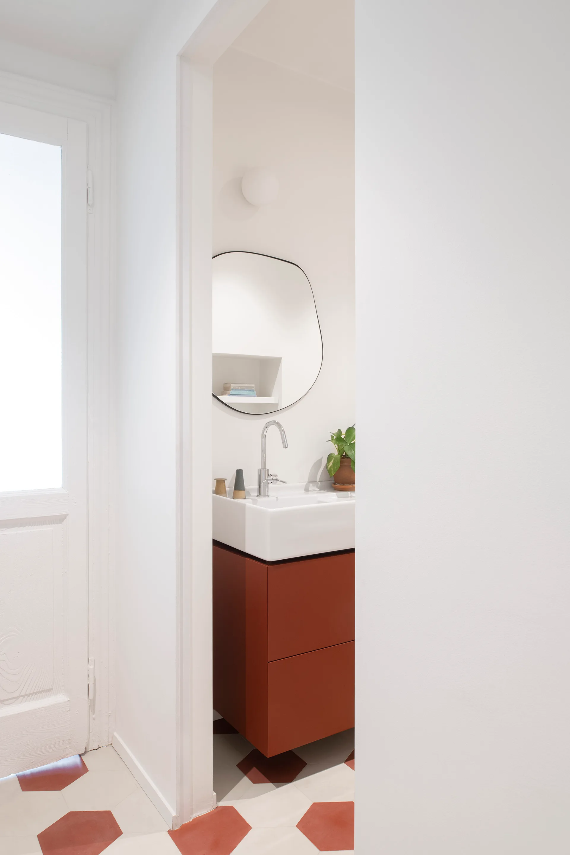 A small bathroom viewed through a doorway features a rust-red floating vanity, an organic-shaped mirror, and hexagon patterned floor tiles.