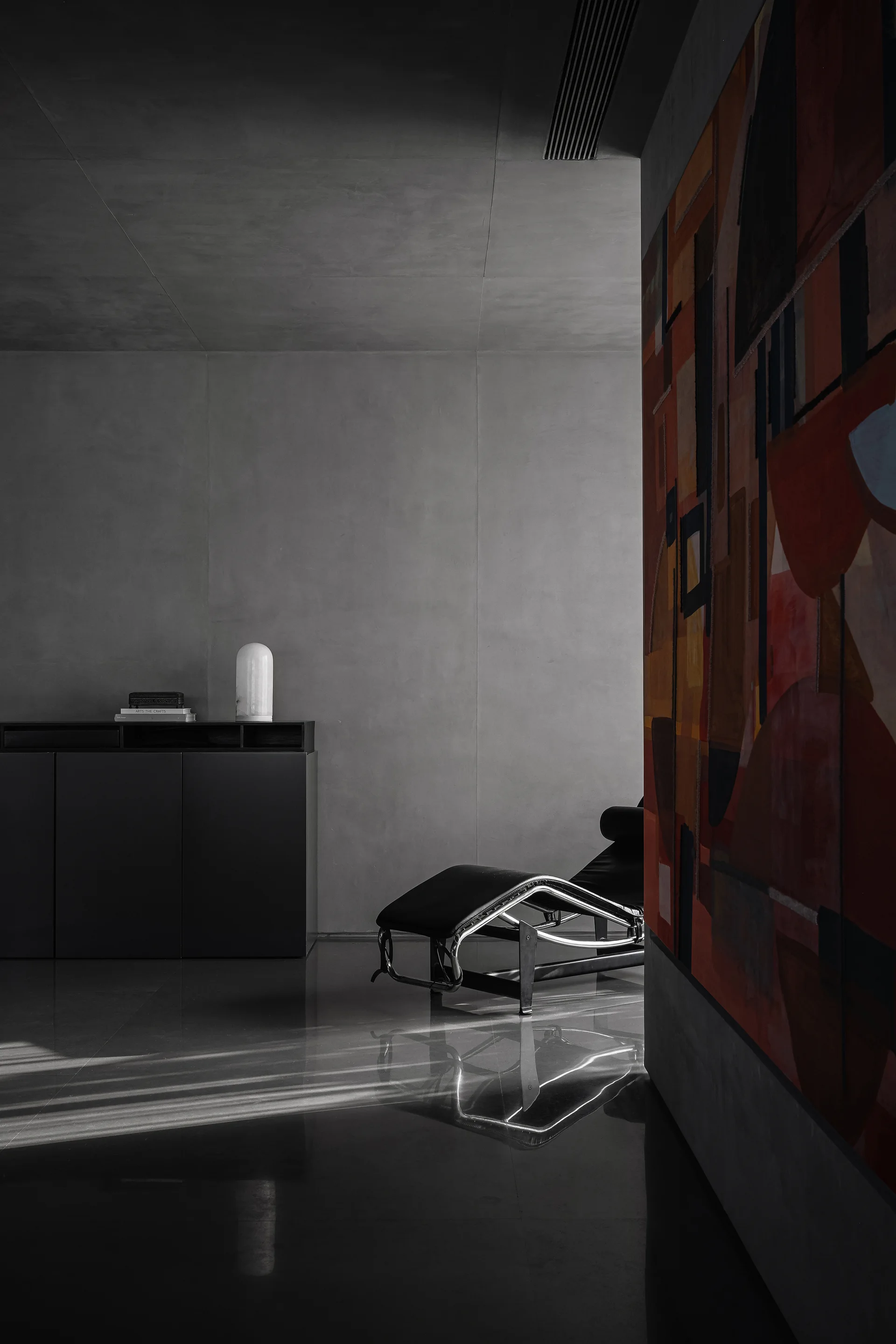 A black leather chaise lounge sits on a polished dark floor in a minimalist gray room next to a low console and a large abstract painting.