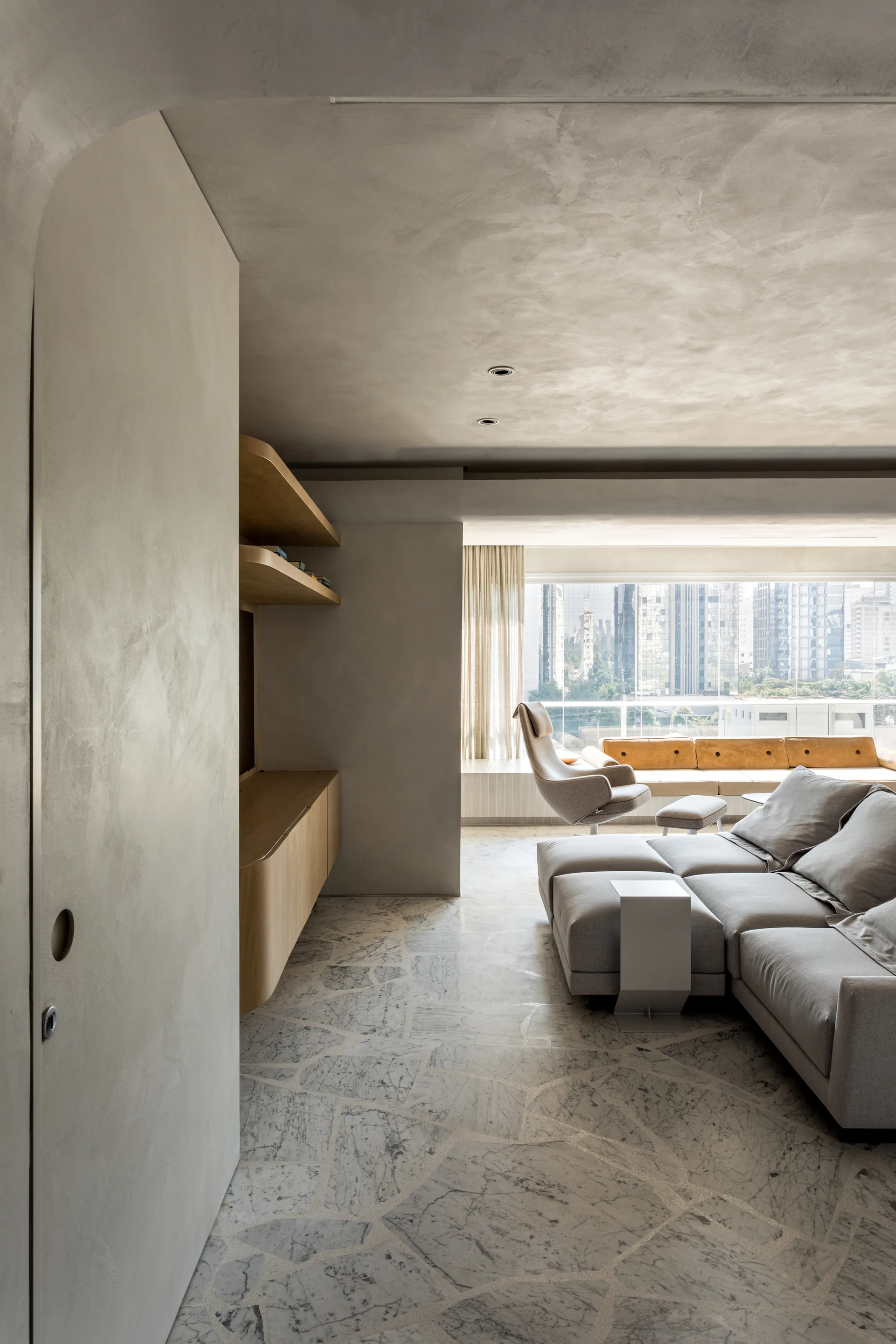 A modern living room features a grey sectional sofa, curved wooden built-in shelving, textured plaster walls, and a fragmented marble tile floor.