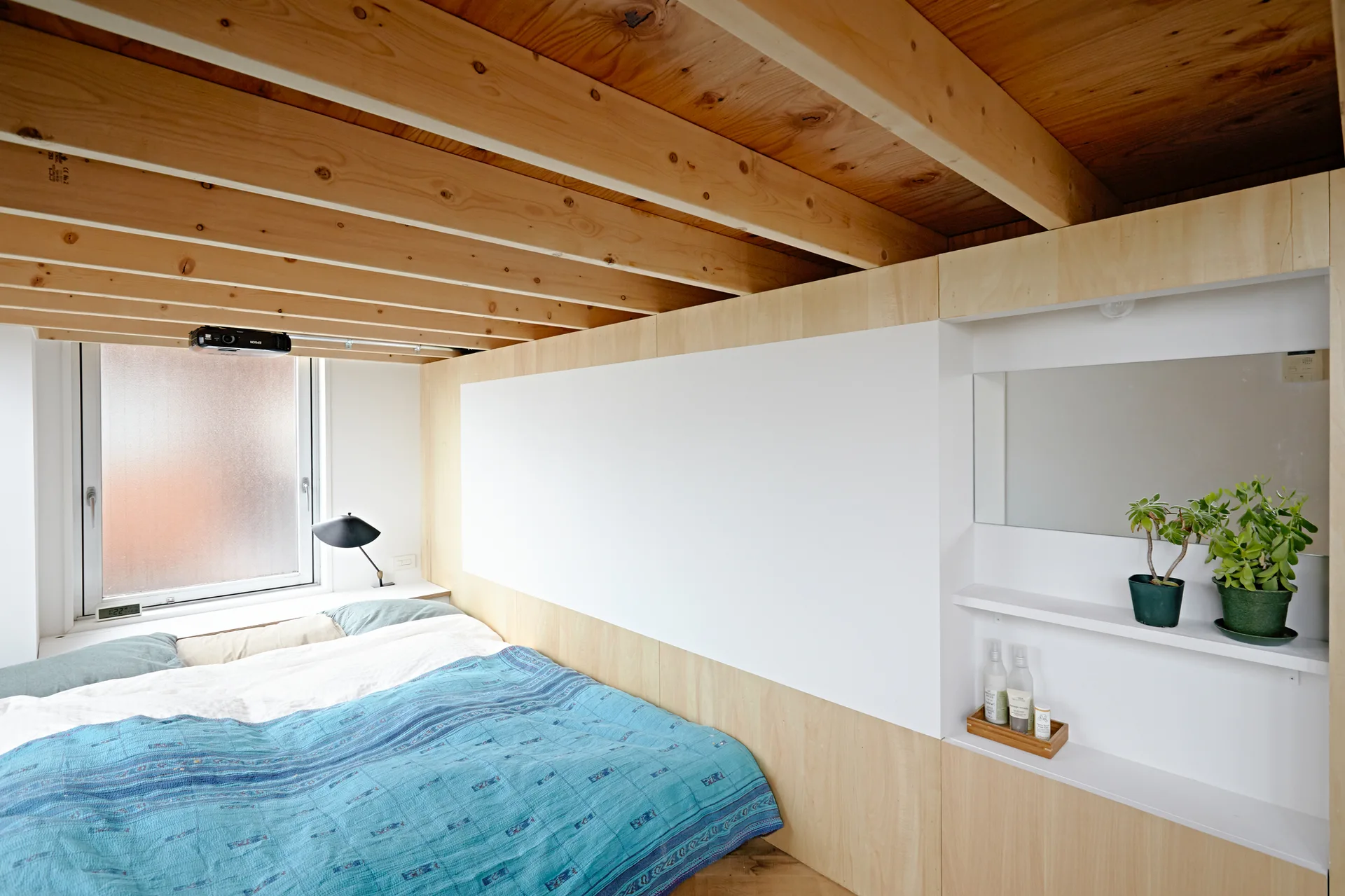 A compact bedroom features a low bed with blue bedding set against a light wood built-in wall storage unit beneath an exposed timber ceiling.