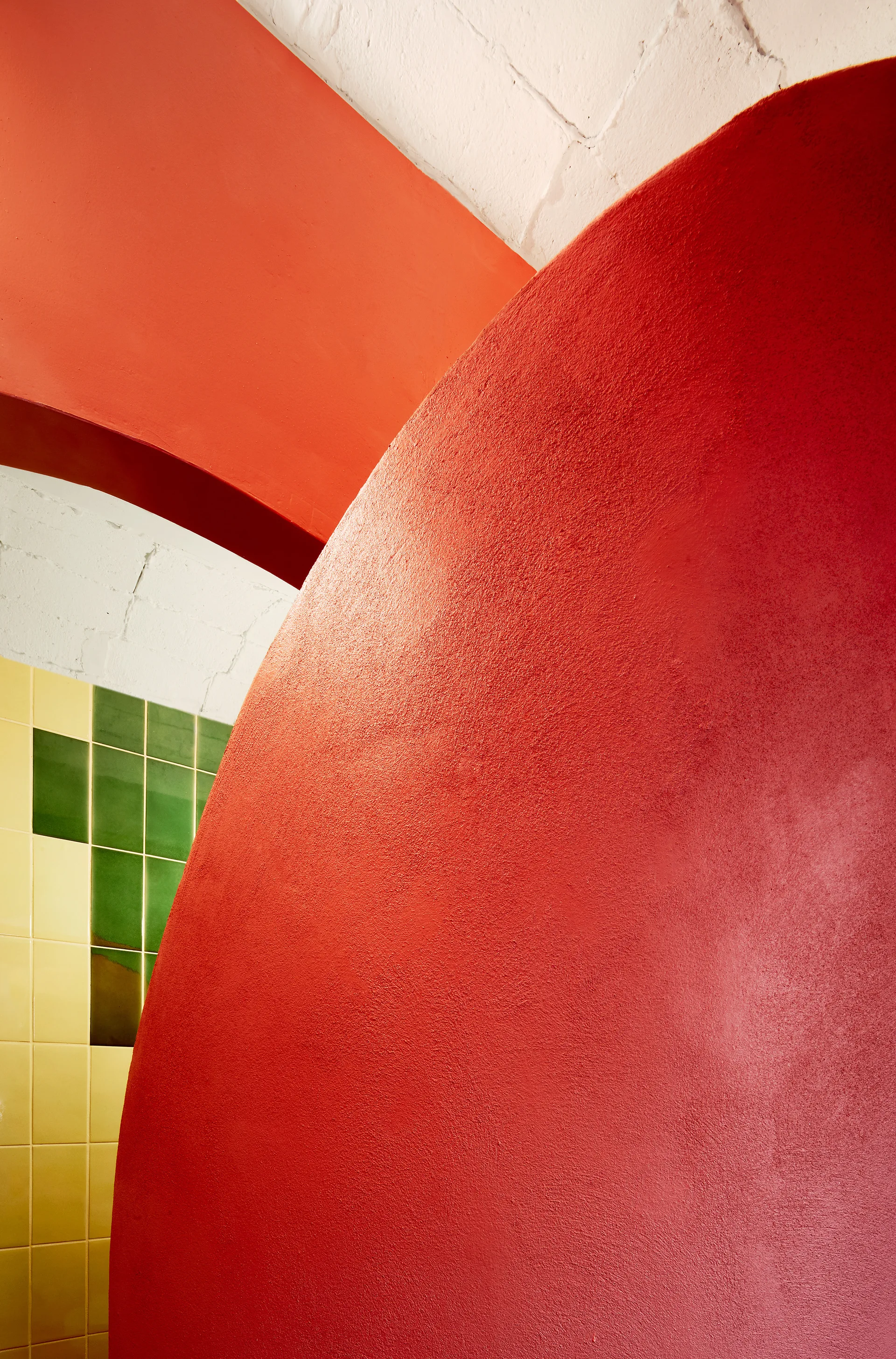 A close-up view of a matte red curved wall meeting a white painted brick ceiling and a background of yellow and green square ceramic tiles.
