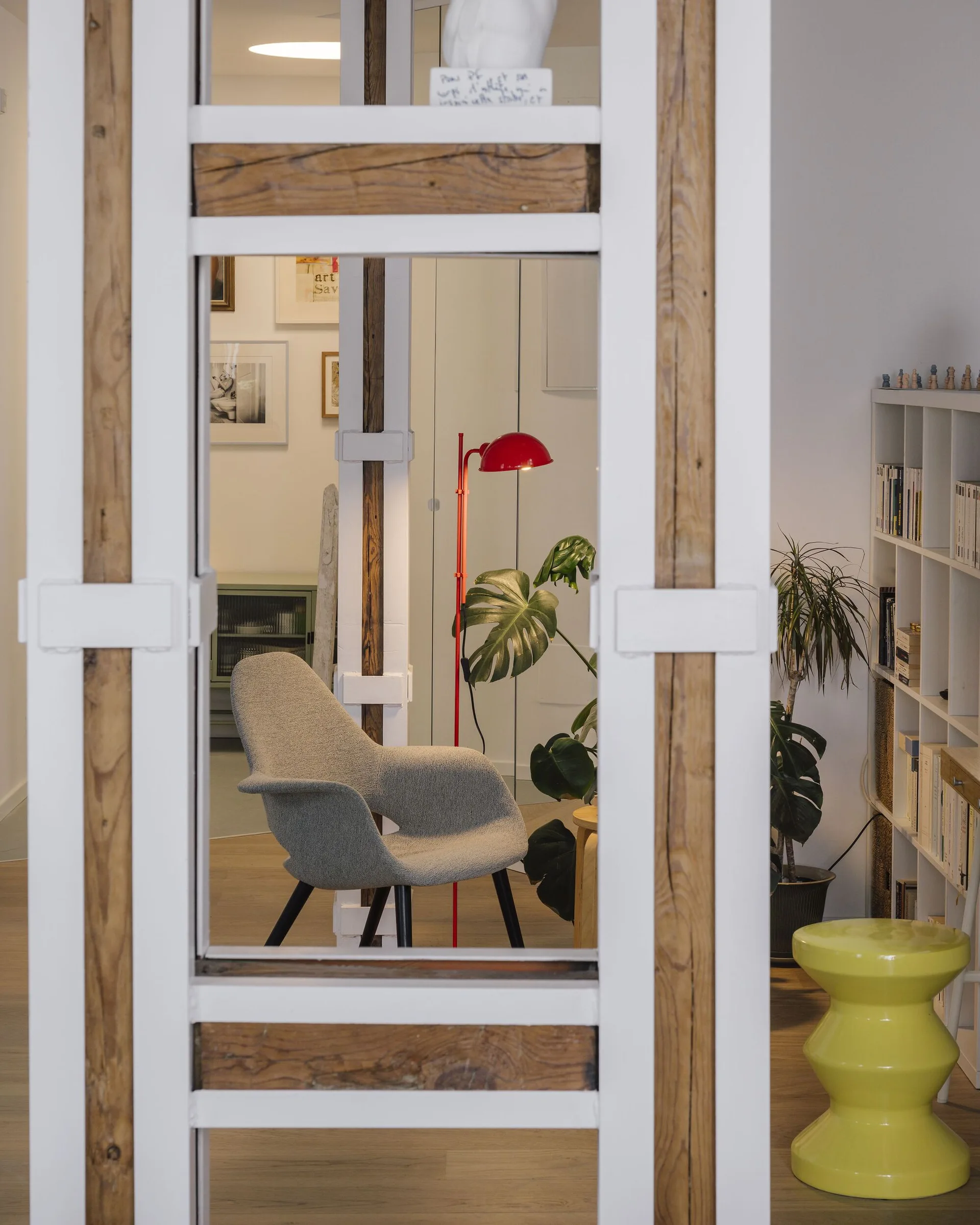 A view through a white and wood architectural frame of a living room featuring a gray armchair, a red floor lamp, and a yellow stool.