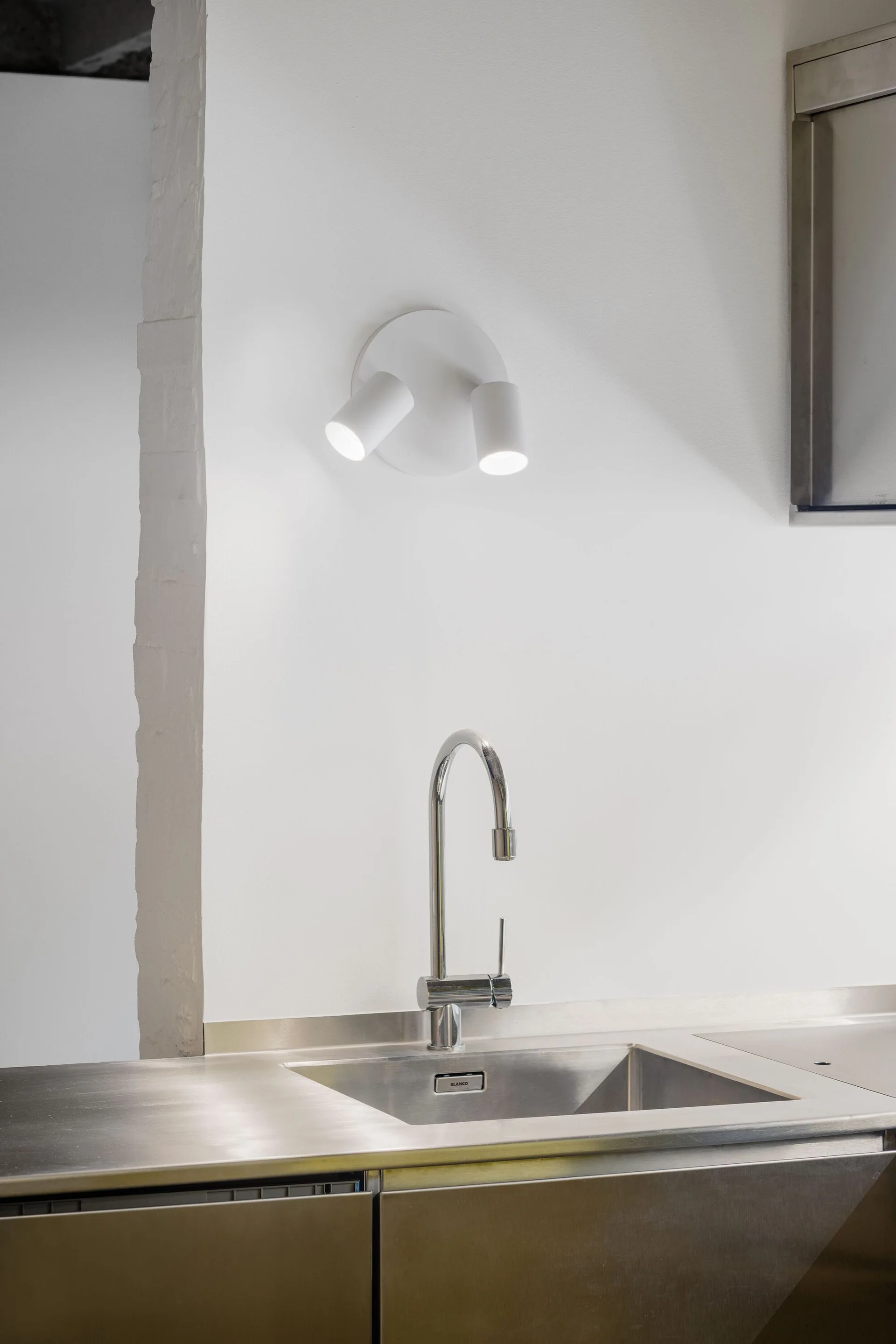 A stainless steel kitchen sink with a chrome gooseneck faucet is integrated into a metal countertop beneath a white wall-mounted dual spotlight.