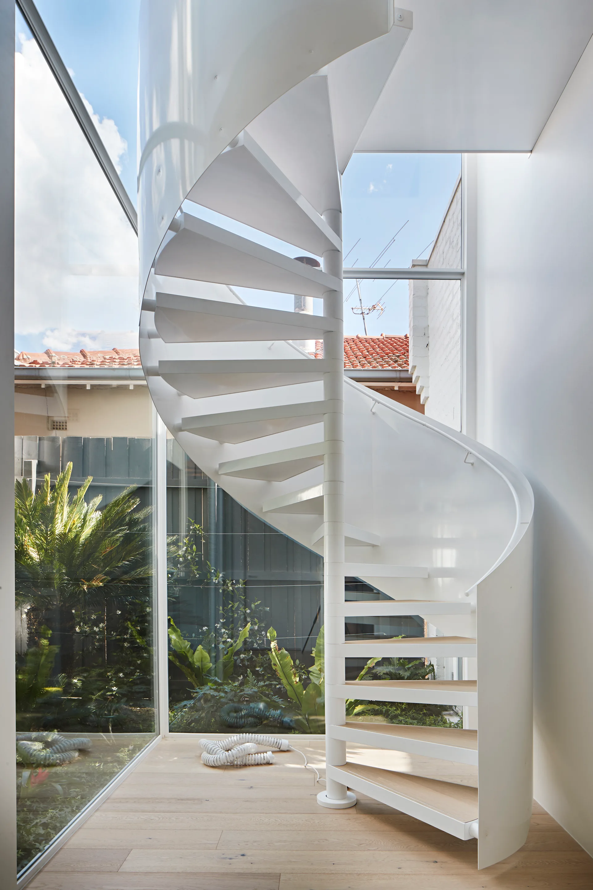 A white metal spiral staircase with light wood steps stands next to floor-to-ceiling glass walls overlooking a garden.