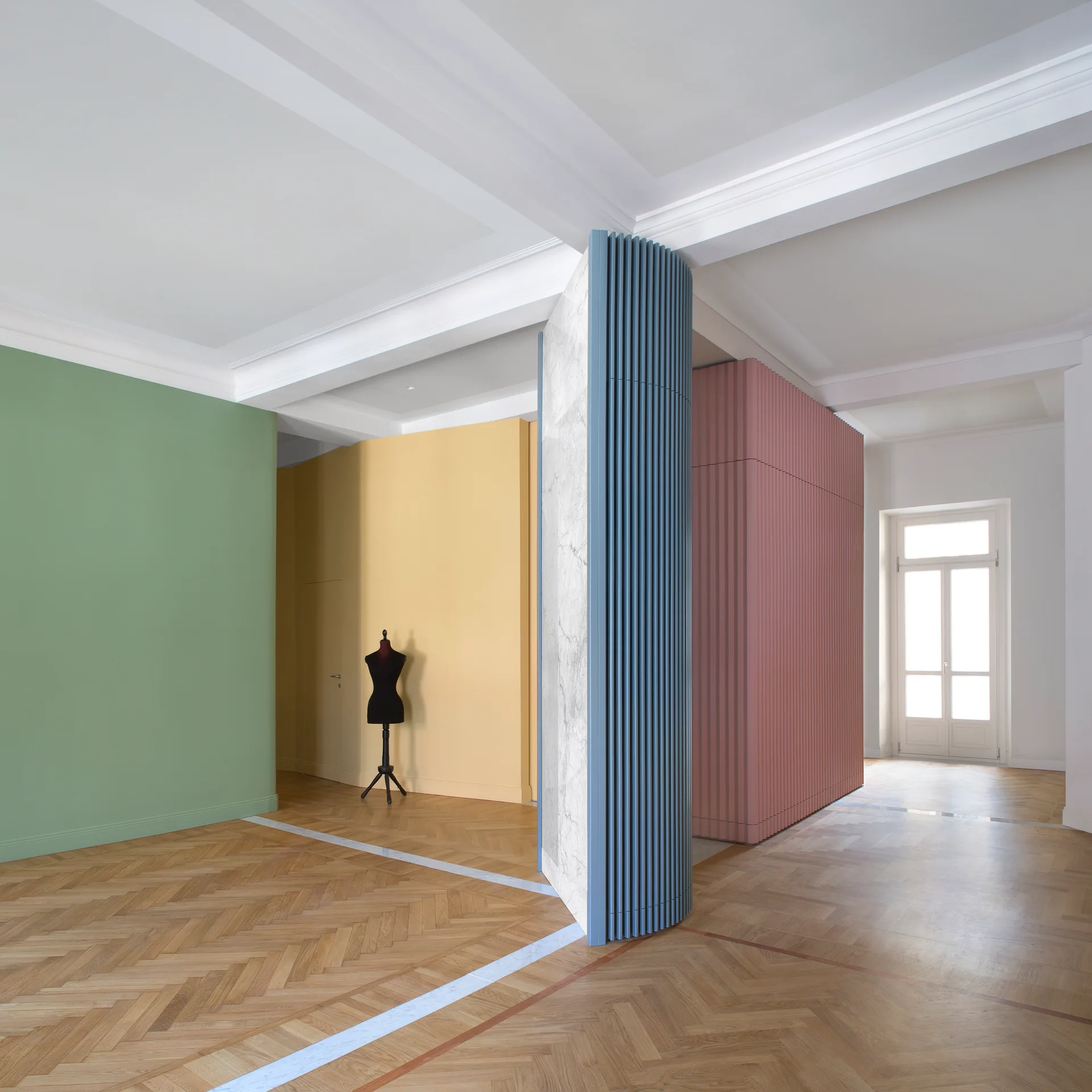A spacious interior features colorful partition walls in yellow, green, blue, and dusty rose alongside a light wood herringbone floor.
