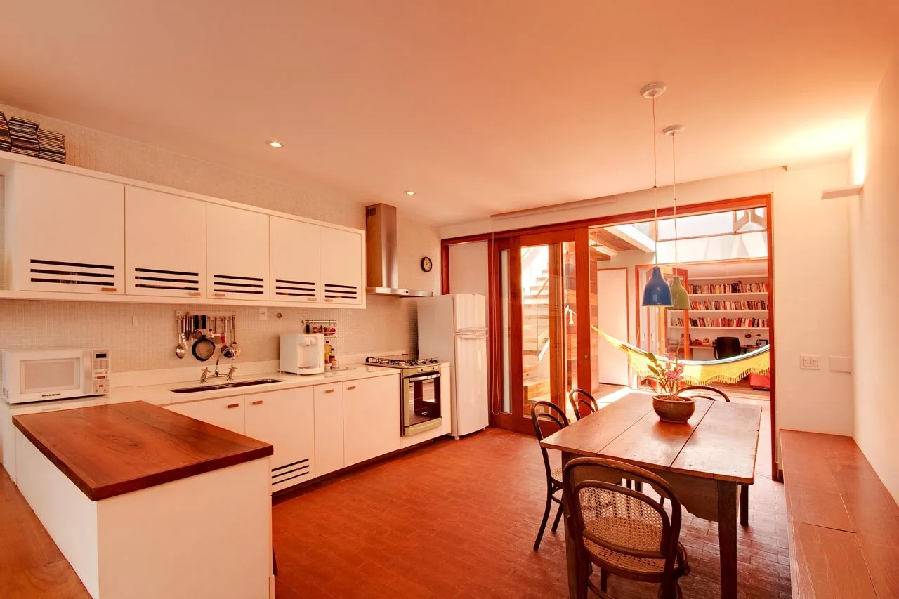 A kitchen with white cabinets and terracotta tile floors features a wooden dining table and a sliding glass door revealing an indoor hammock.