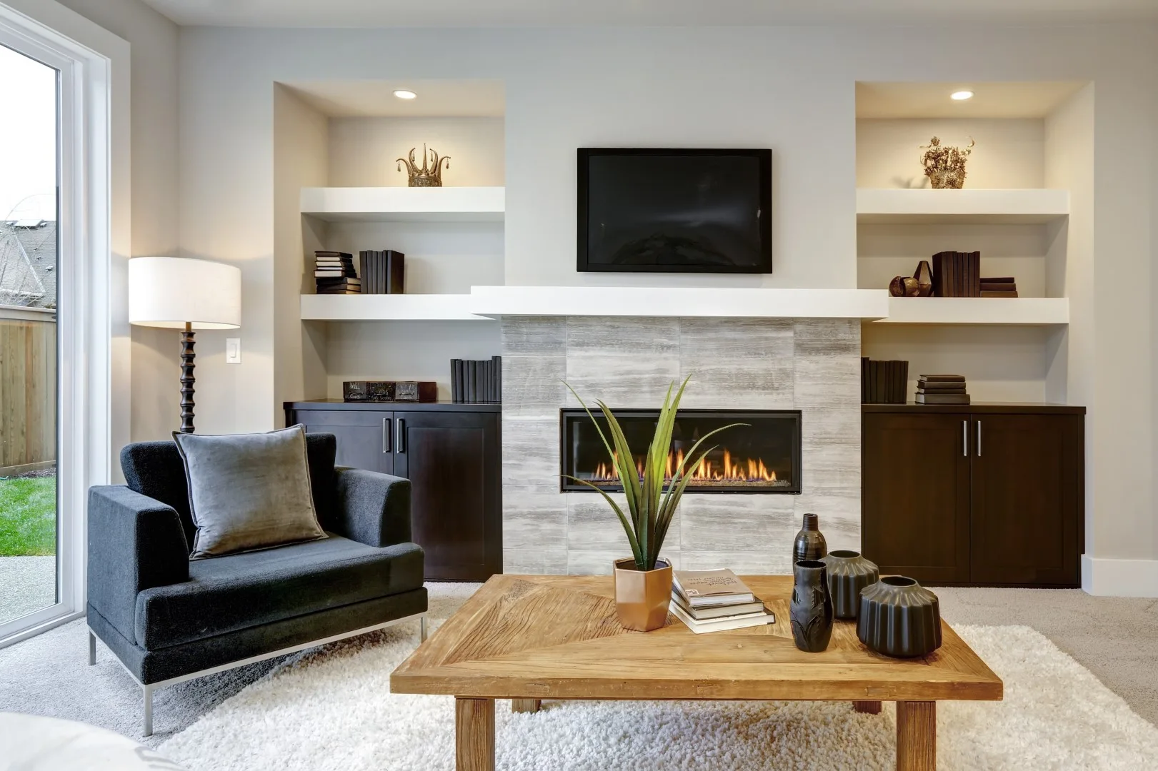 A living room features a tiled fireplace flanked by recessed white shelves, dark lower cabinets, a black armchair, and a rustic wooden coffee table.