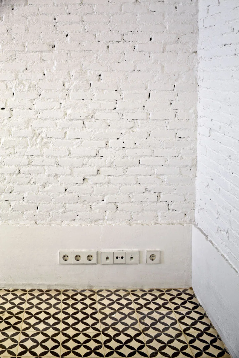 A white painted brick wall meets black and white geometric patterned floor tiles in a minimalist room corner with integrated electrical wall outlets.