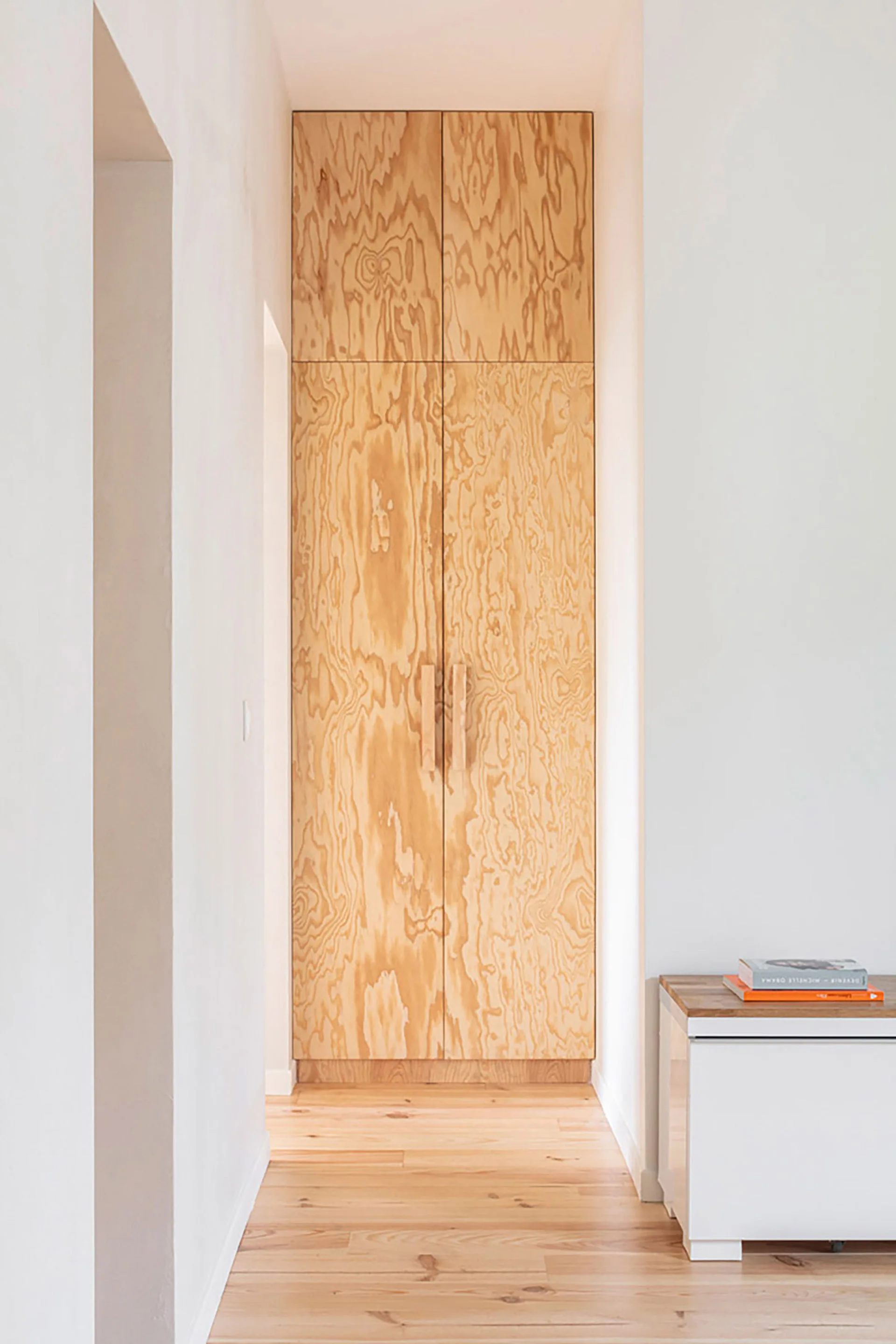 A tall floor to ceiling plywood cabinet stands at the end of a white hallway with light wood floors and a low white cabinet.