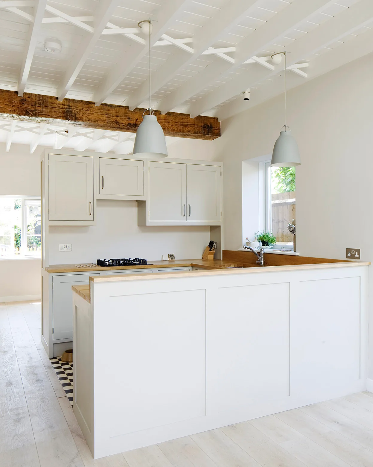 Cream-colored shaker cabinetry and wood countertops define a kitchen with gray pendant lights, a checkered tile floor, and an exposed wooden beam.