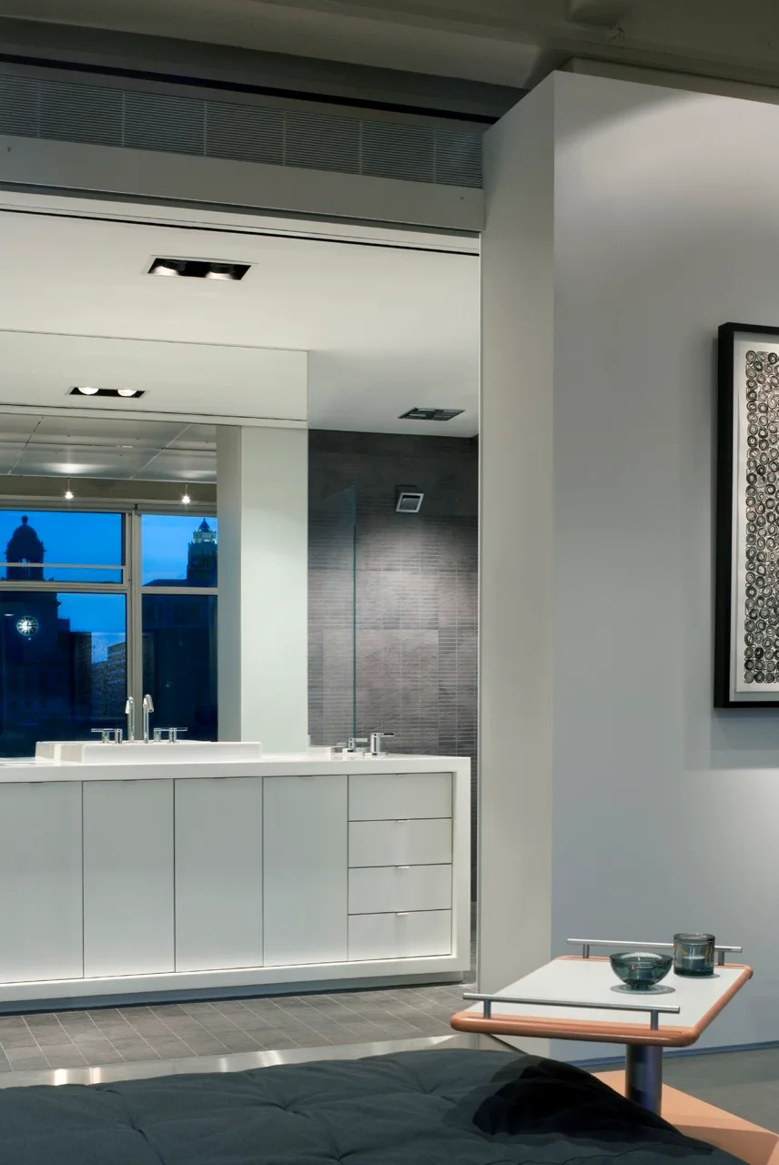 A minimalist white vanity features a sleek chrome faucet and a large mirror reflecting a dark tile shower wall and a city skyline at dusk.