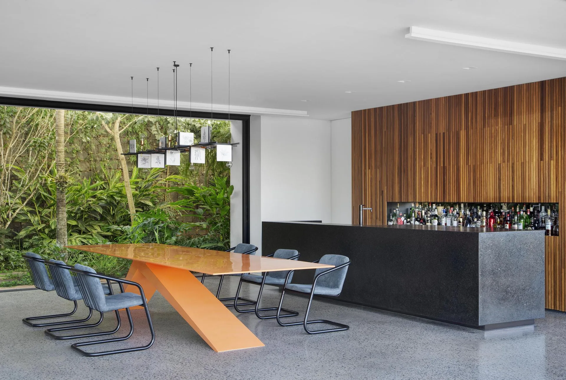 A modern dining room features an angular orange table, blue chairs, a black stone bar, and a vertical wood-slat wall opening to a garden.