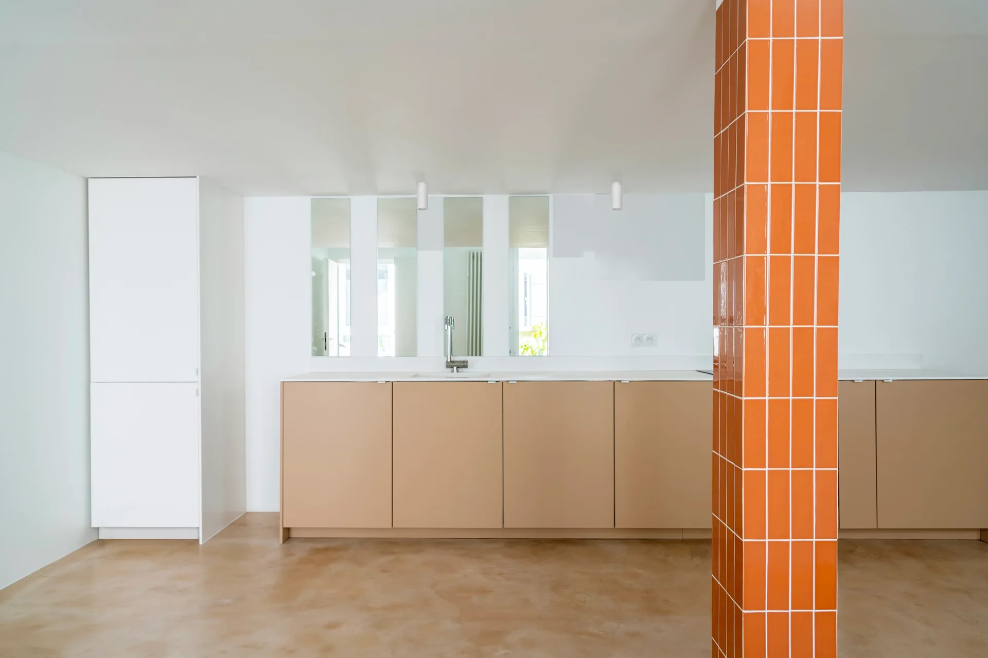 A minimalist kitchen features flat-panel beige cabinets, vertical strip mirrors, and a prominent structural pillar clad in glossy orange tiles.