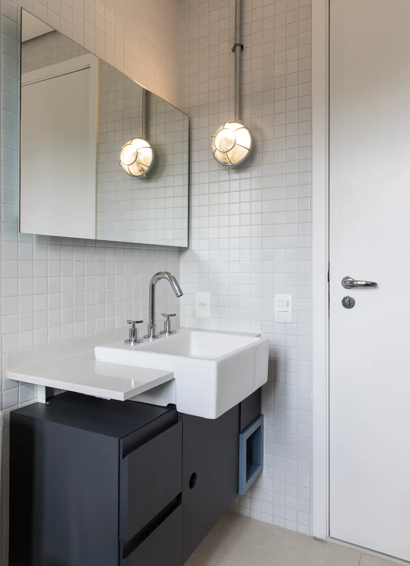 A dark gray bathroom vanity with a white sink sits against white square grid wall tiles below a large mirror and an industrial wall sconce.