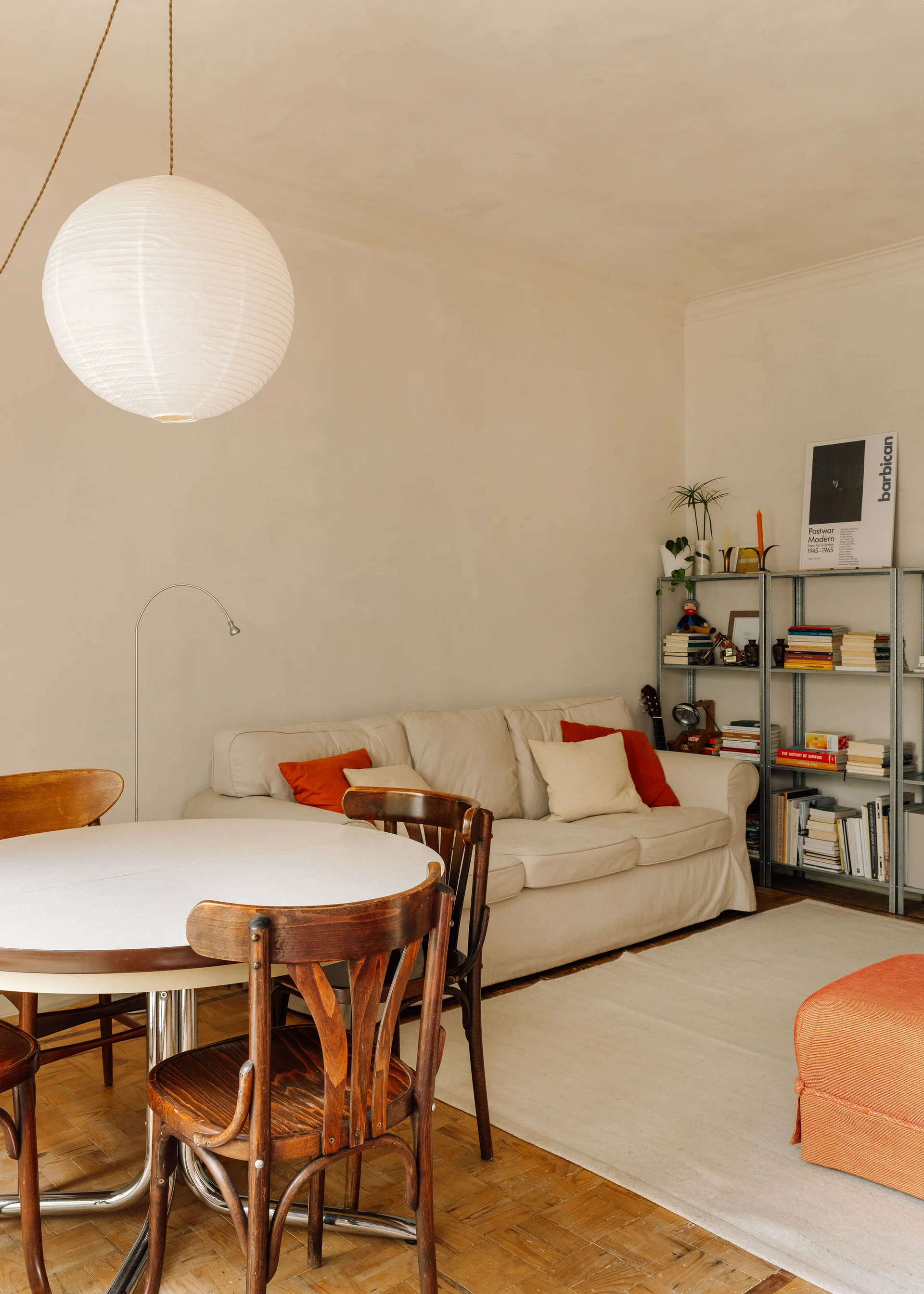 A cream sofa with red accent pillows sits near a round wooden dining table with bentwood chairs under a large paper globe pendant light.