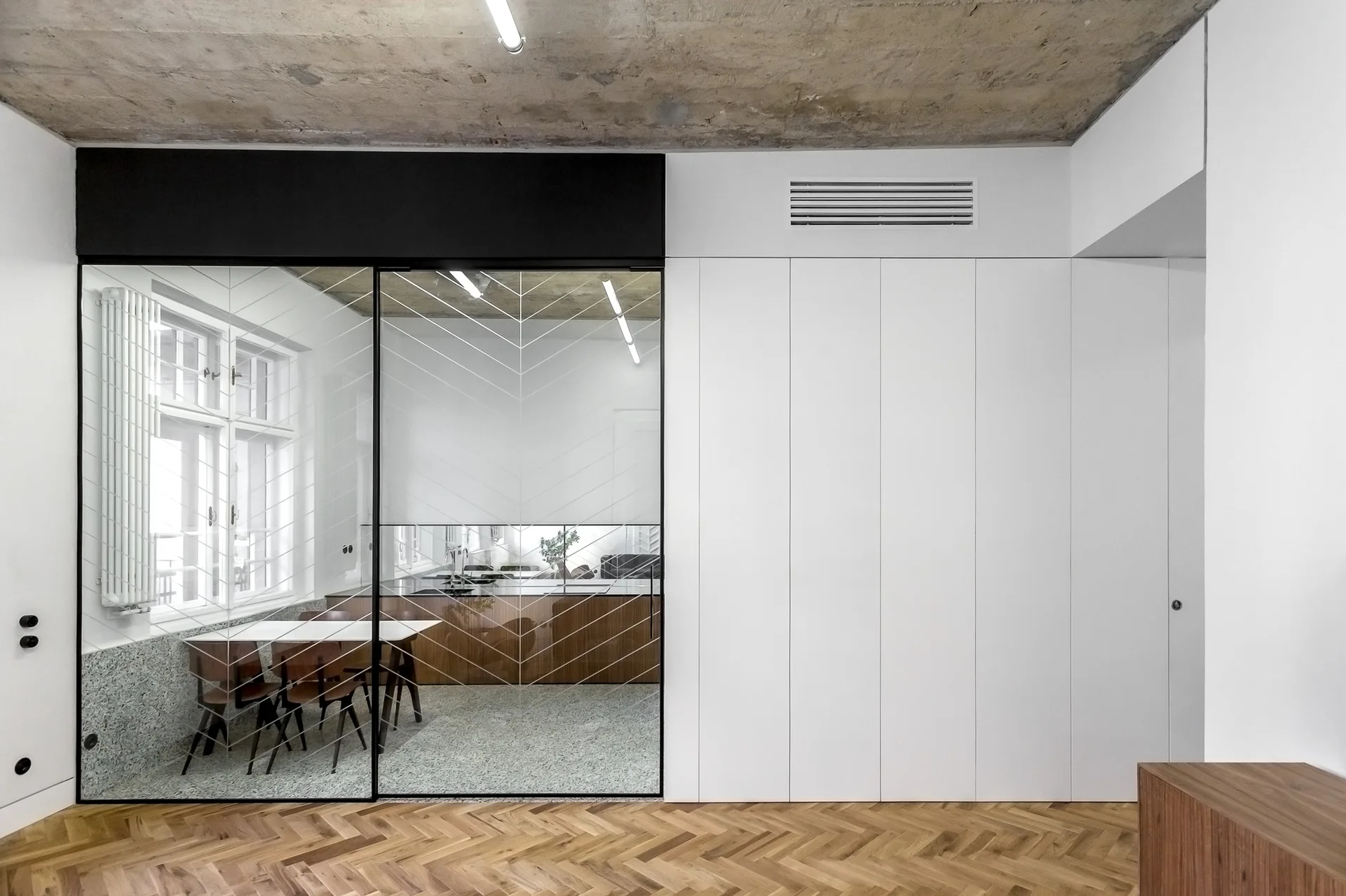 A minimalist room featuring a chevron-patterned glass partition, flat-panel white cabinets, a raw concrete ceiling, and herringbone wood floors.