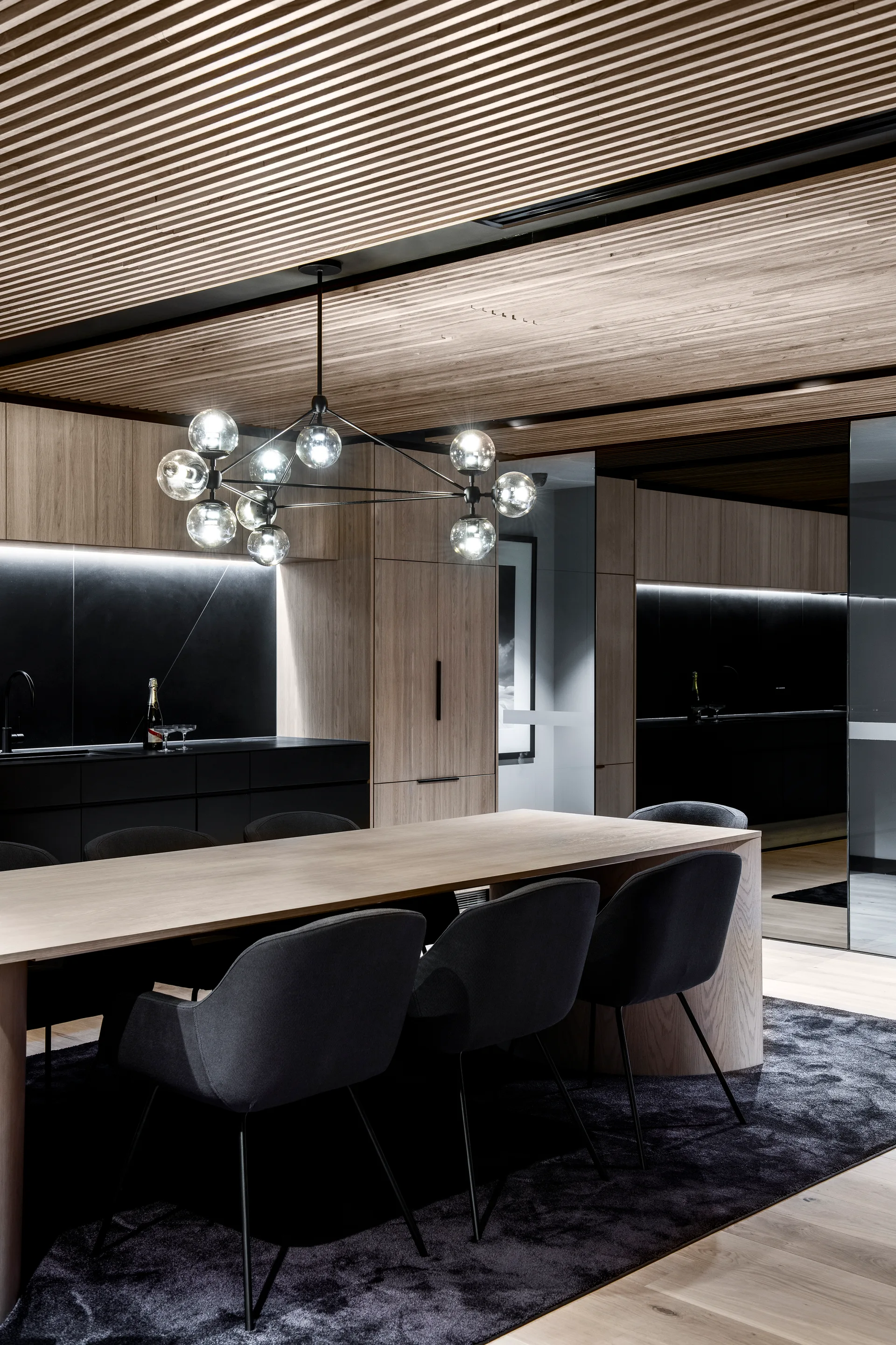 A modern dining room features a light wood table, charcoal chairs, a branching globe chandelier, and a slatted wood ceiling.