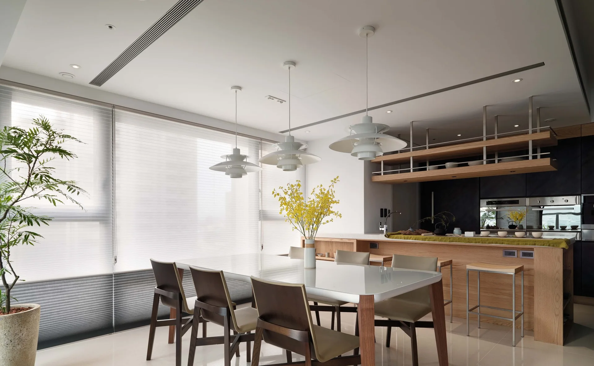 A modern dining area features a glass-topped table, dark wood chairs, three white pendant lights, and a wooden kitchen island in the background.