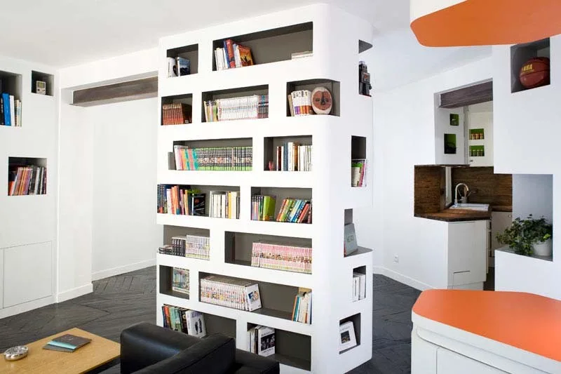 A modern white modular room divider bookshelf sits on dark herringbone wood flooring next to a kitchen.