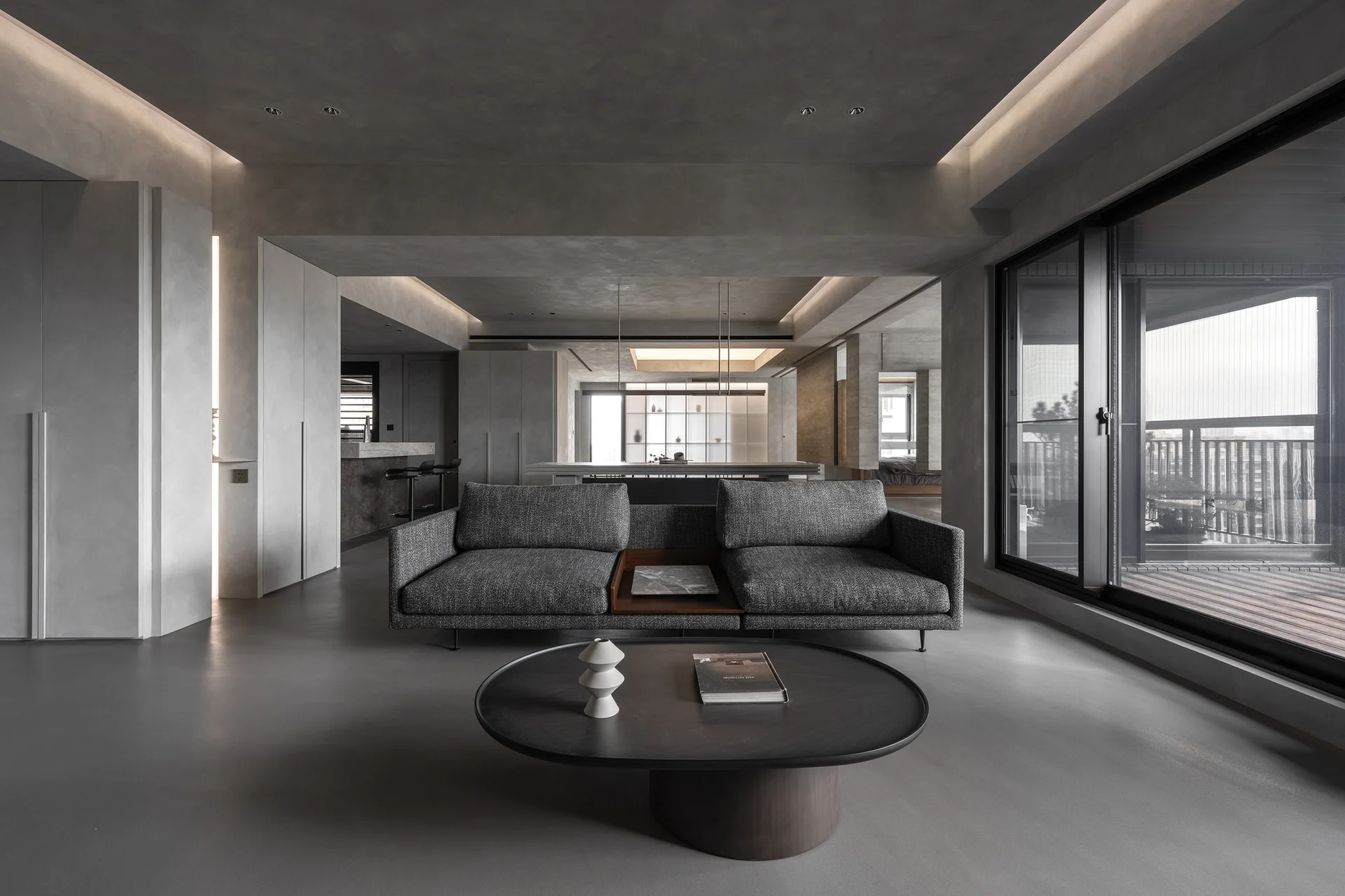 A minimalist grey living room features a dark grey fabric sofa, a dark round coffee table, and stepped ceiling cove lighting.