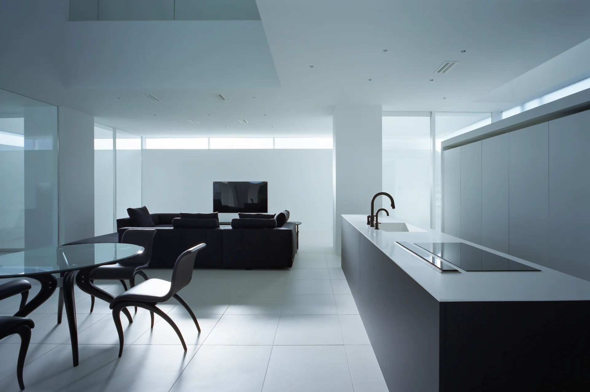 A minimalist open-concept room features a black kitchen island with curved faucet, a glass-top dining table, and a dark sectional sofa on white tile floors.