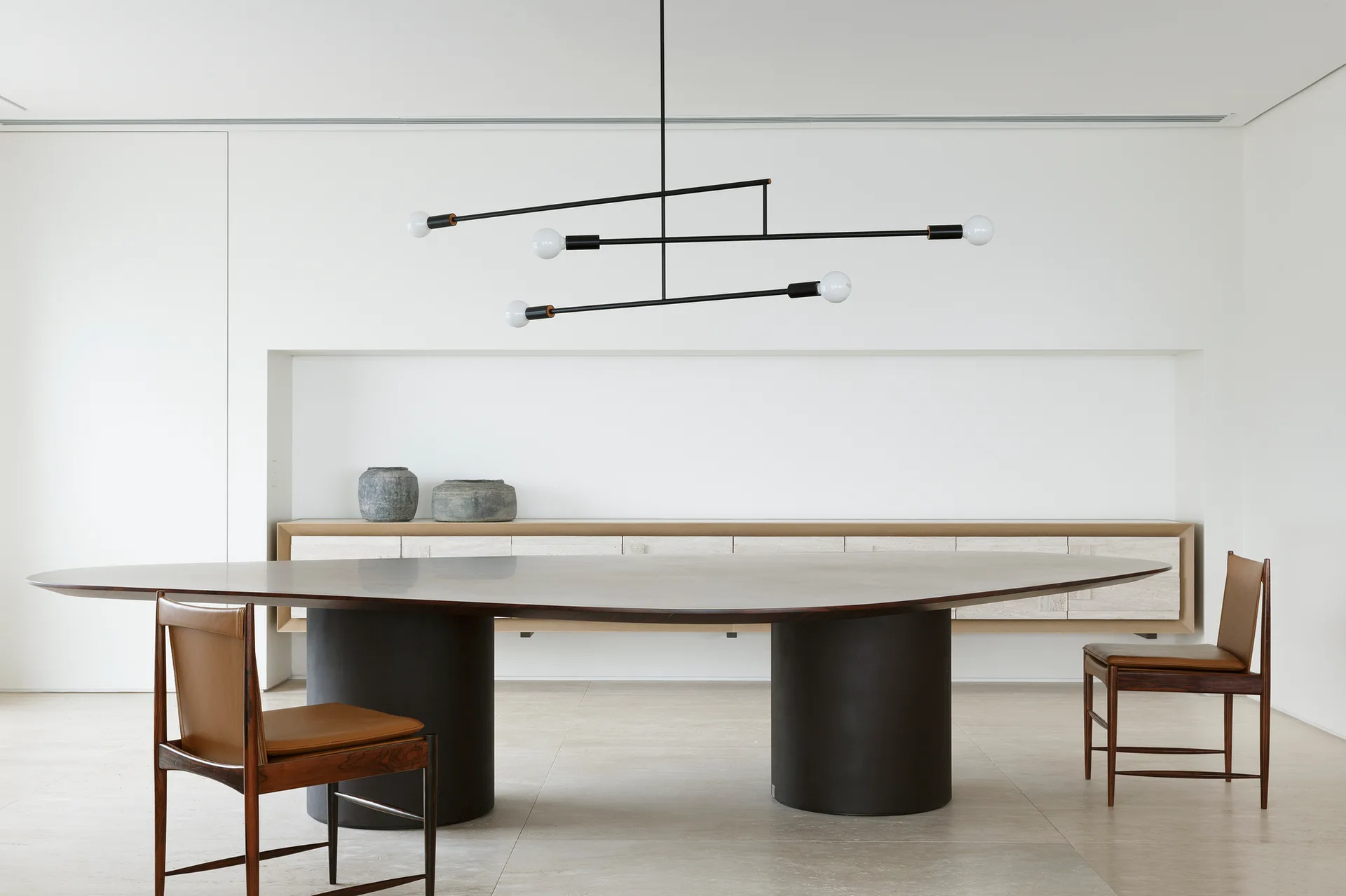 A minimalist dining room features a dark oval table on pedestal bases, two leather chairs, a black linear pendant light, and a built-in stone credenza.