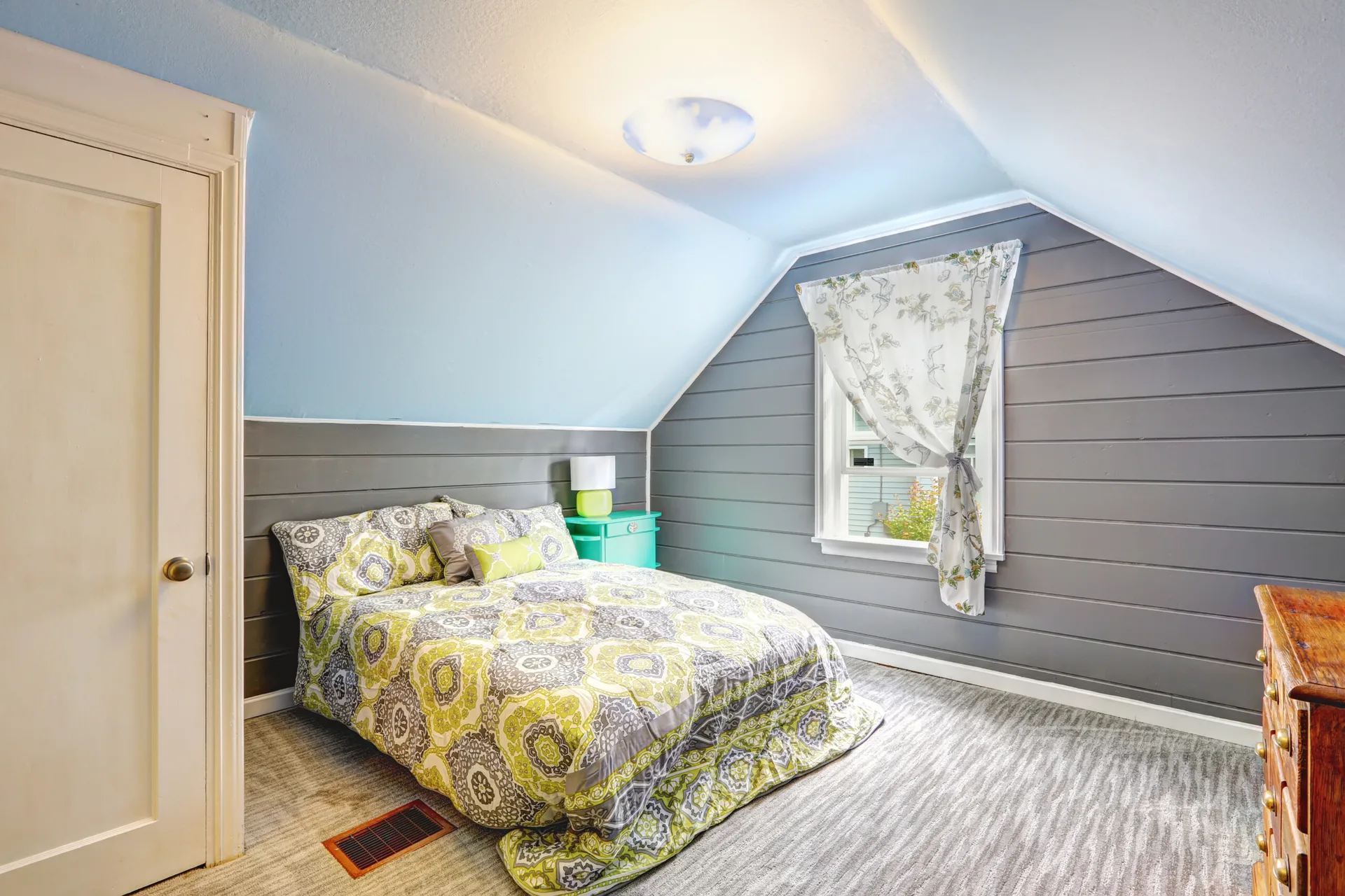 A small attic bedroom featuring grey horizontal plank walls, a sloped light blue ceiling, a bed with green patterned bedding, and a white door.