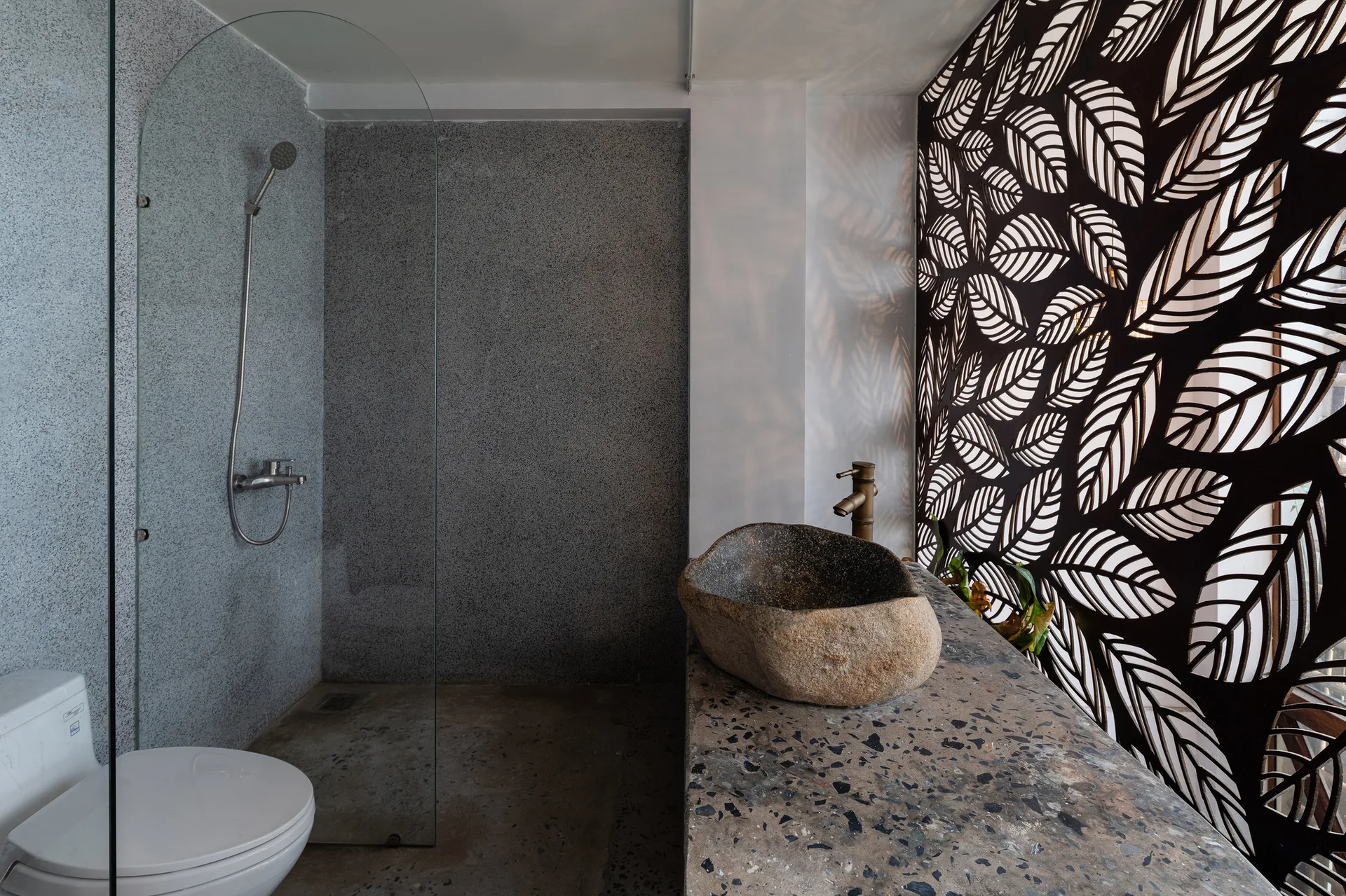 A modern bathroom features a rough stone vessel sink on a terrazzo vanity next to a decorative laser-cut leaf pattern screen.