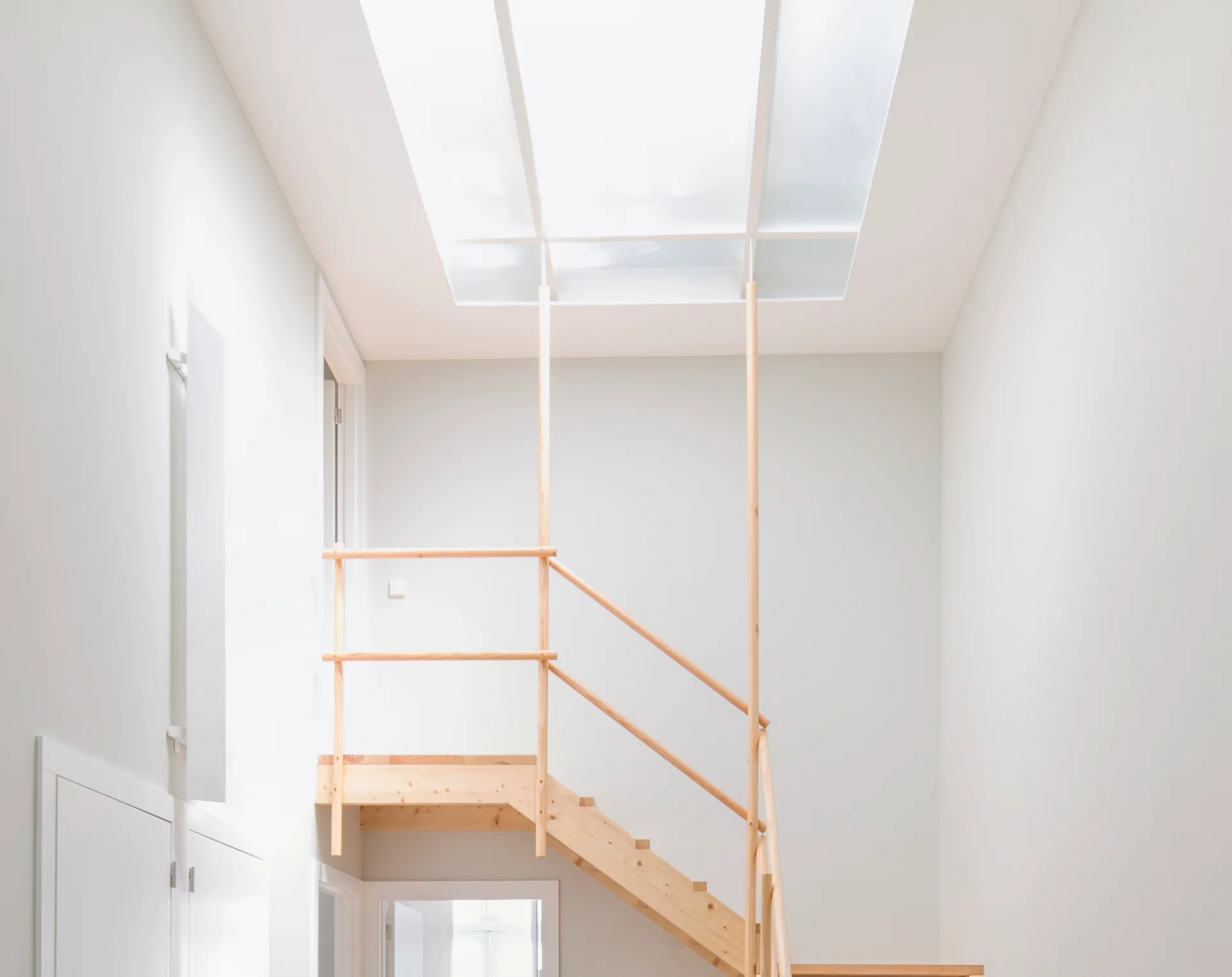 A minimalist white stairwell features a light wood staircase with a simple wood handrail, illuminated by a large rectangular skylight overhead.