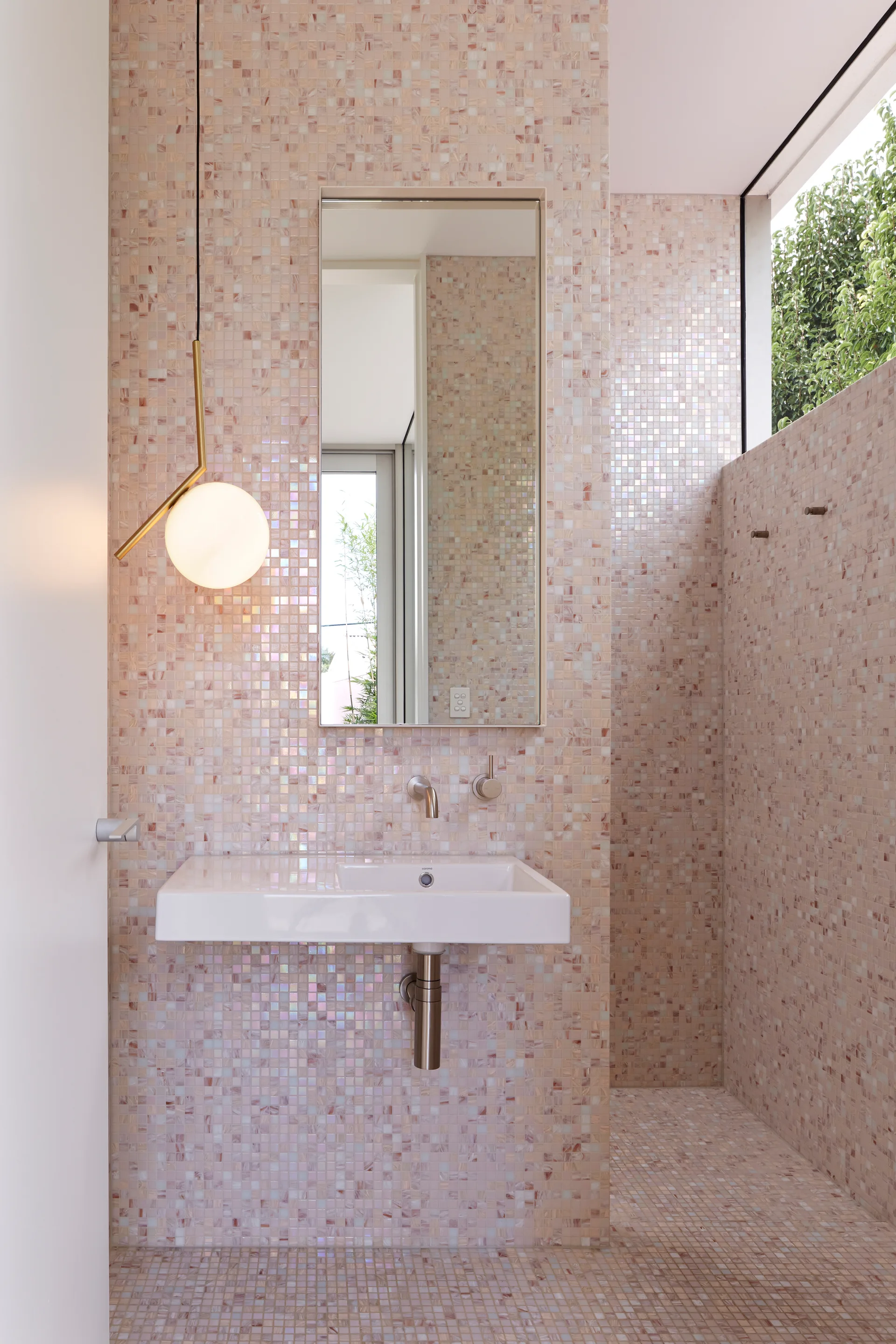 A minimalist bathroom with pink iridescent mosaic tiled walls, a white wall-mounted sink, a rectangular mirror, and a gold globe pendant light.