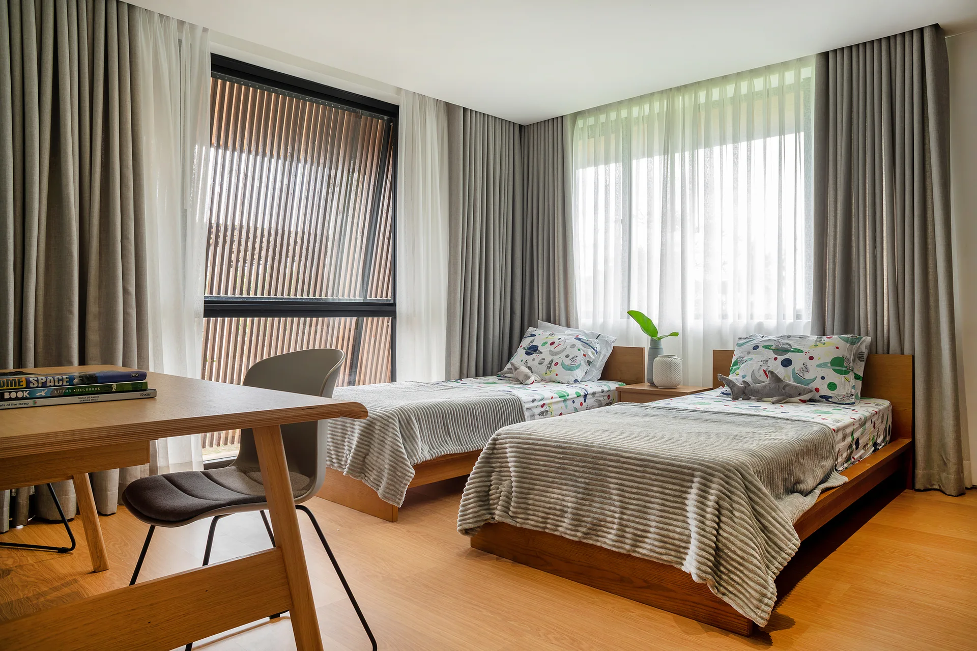 A shared children's bedroom featuring two wooden twin beds with space-themed sheets, a desk, and floor-to-ceiling windows with layered grey curtains.