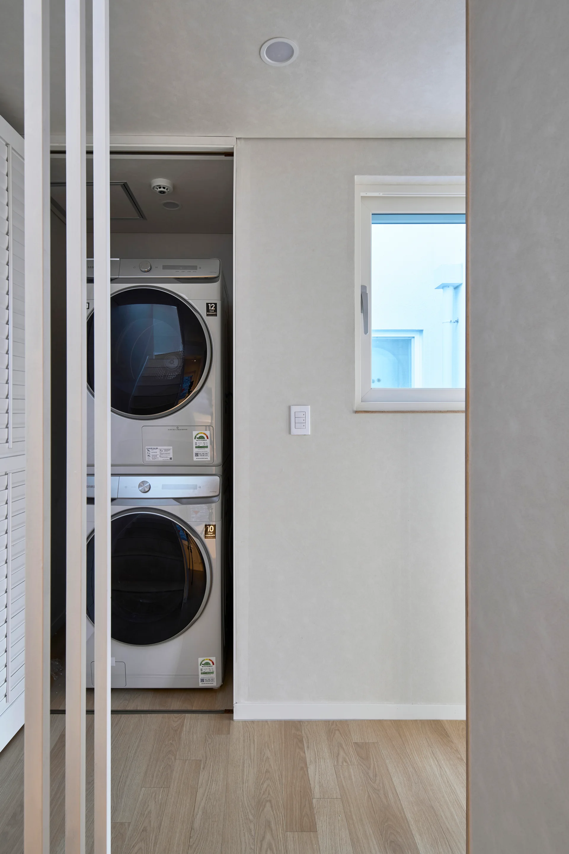 A compact utility area features a stacked silver washer and dryer set behind a white slatted partition wall next to a small window.