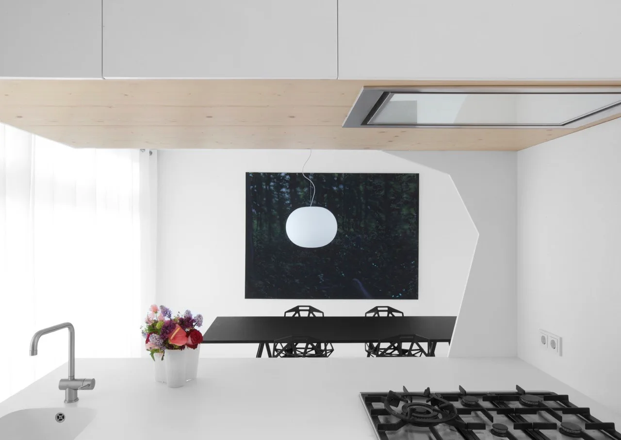 A minimalist white kitchen island with a gas stovetop and faucet looks through a geometric cutout wall to a dining area with black table and chairs.