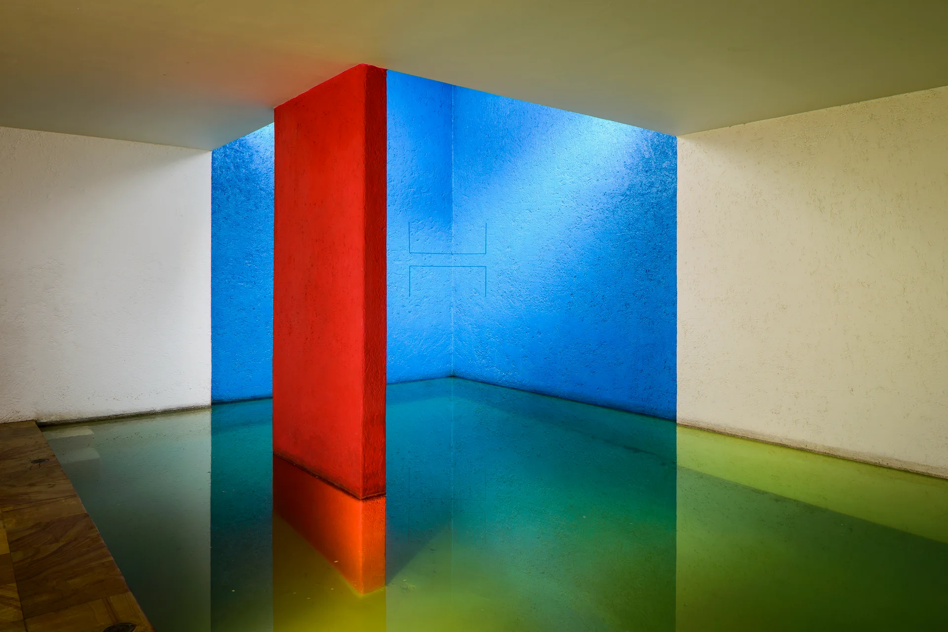 A minimalist indoor pool with a reflective water surface surrounded by textured blue, white, and red walls under natural skylight.