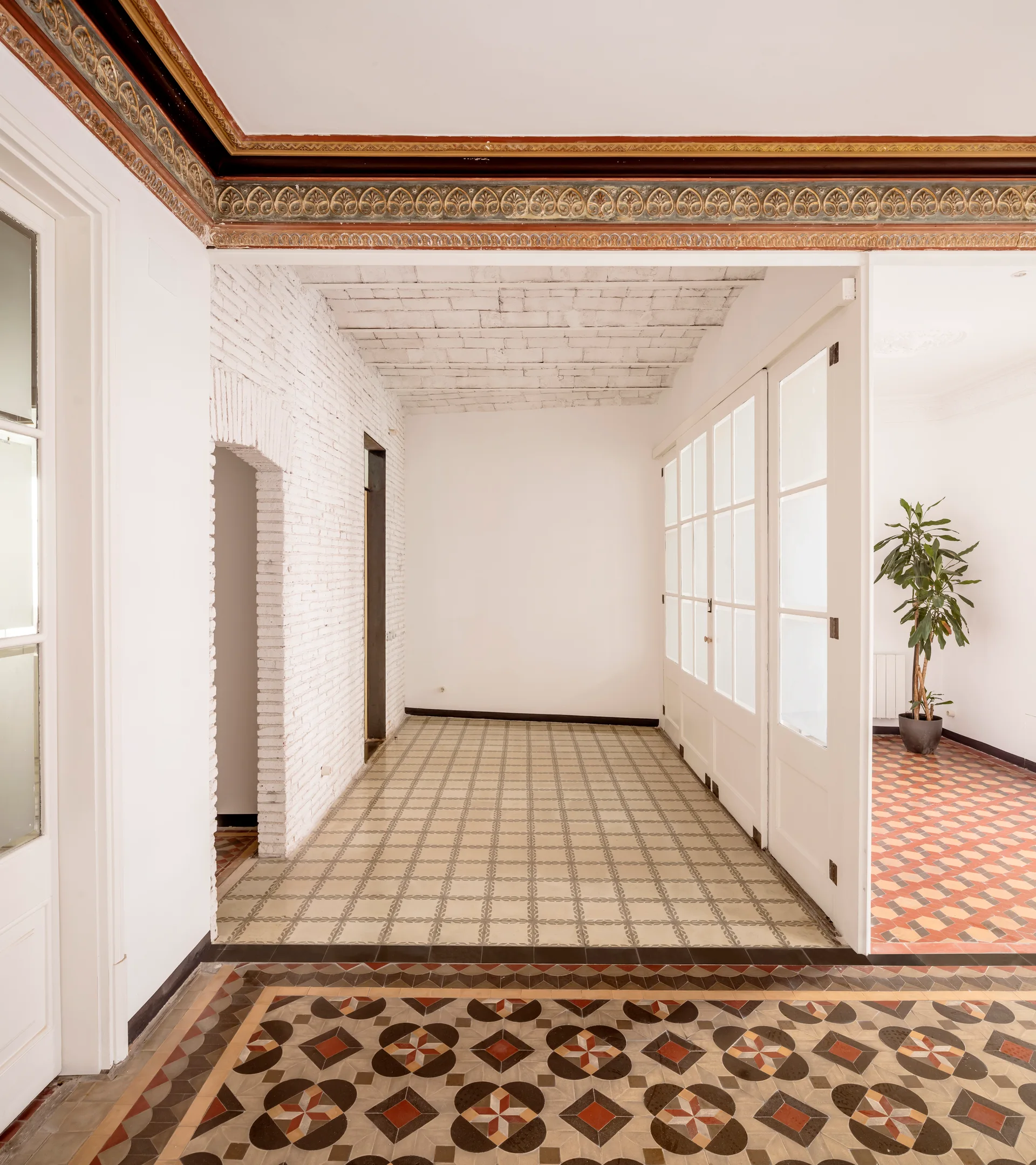 A bright interior hallway featuring geometric patterned tile flooring, a white-painted brick wall, and ornate colorful ceiling molding.