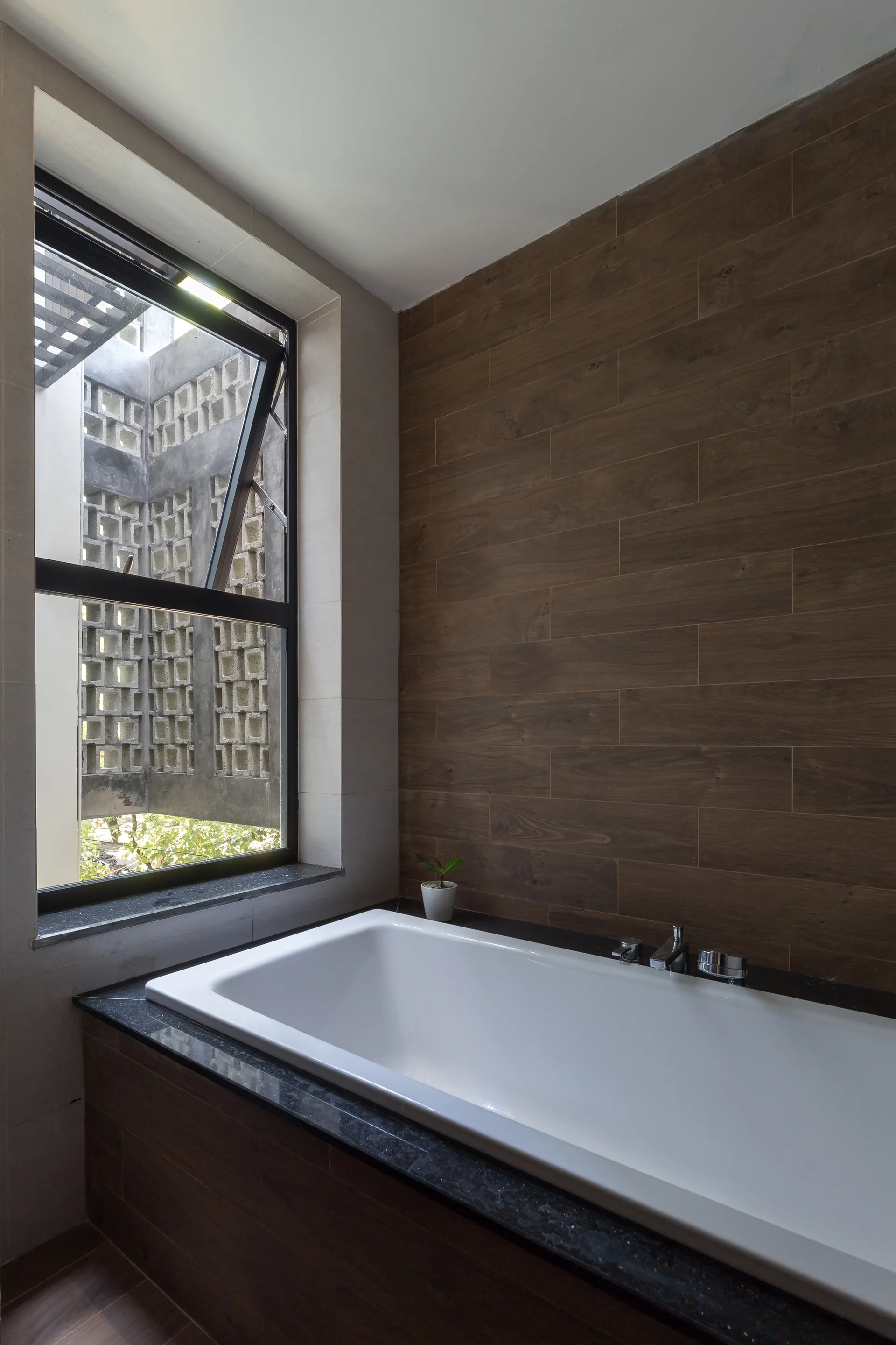 A modern bathroom features a white built-in bathtub surrounded by black stone, next to a wood-look tile wall and a window showing a concrete block screen.