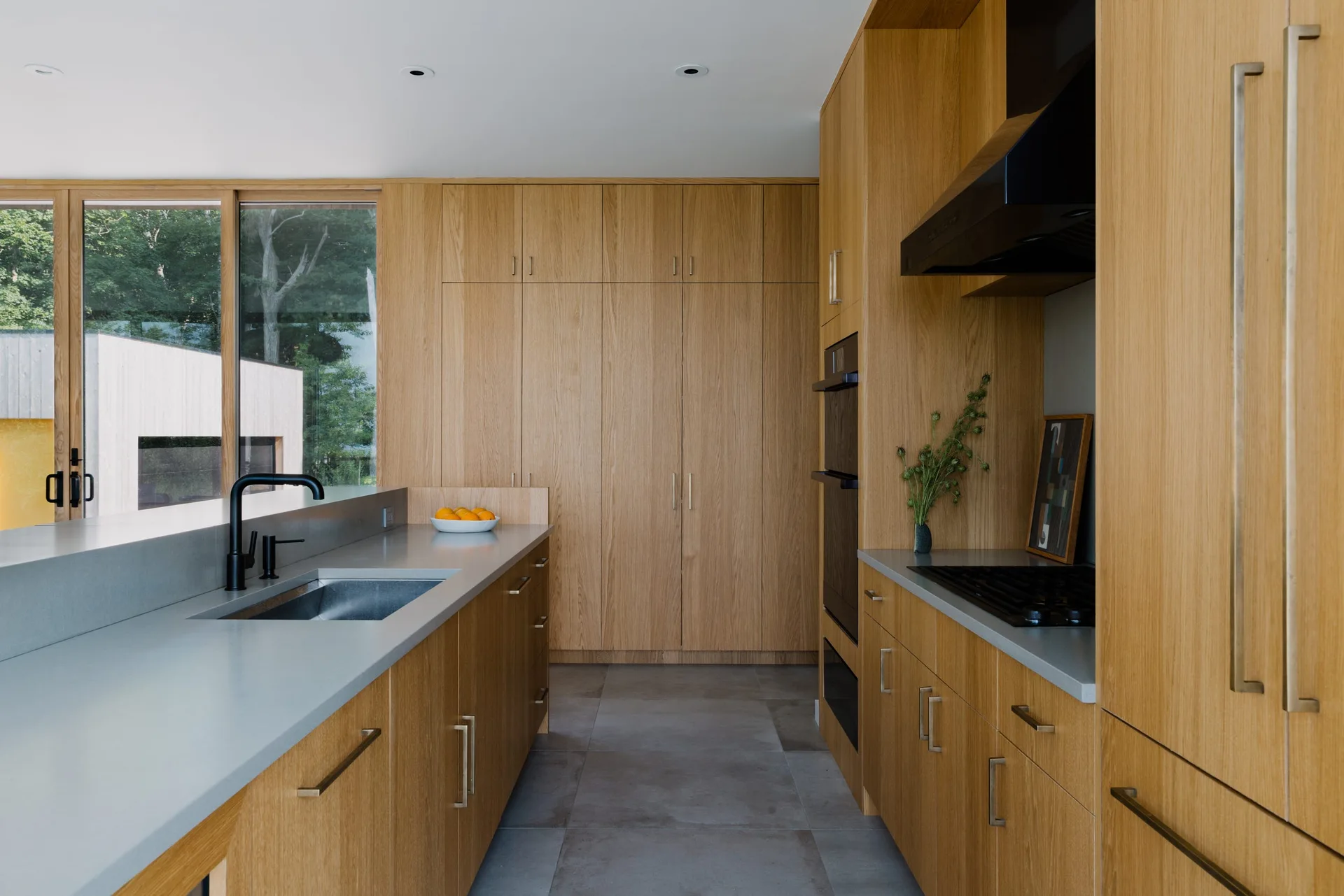 A minimalist kitchen features full-height light wood cabinets, matte gray countertops with an integrated sink, and large format gray floor tiles.