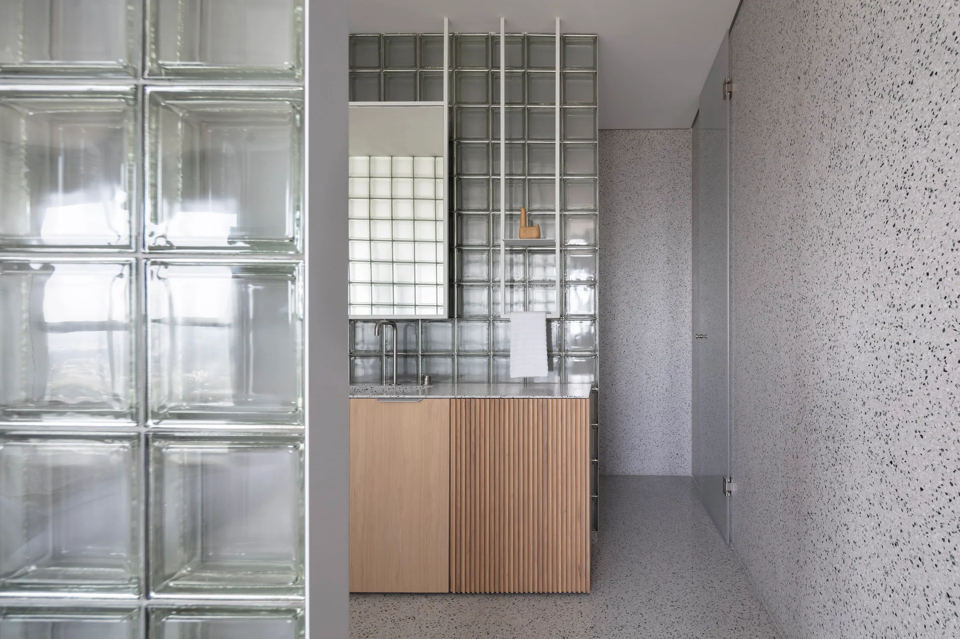 A minimalist bathroom features speckled grey terrazzo walls, a glass block partition, and a light wood vanity with fluted and flat cabinet doors.