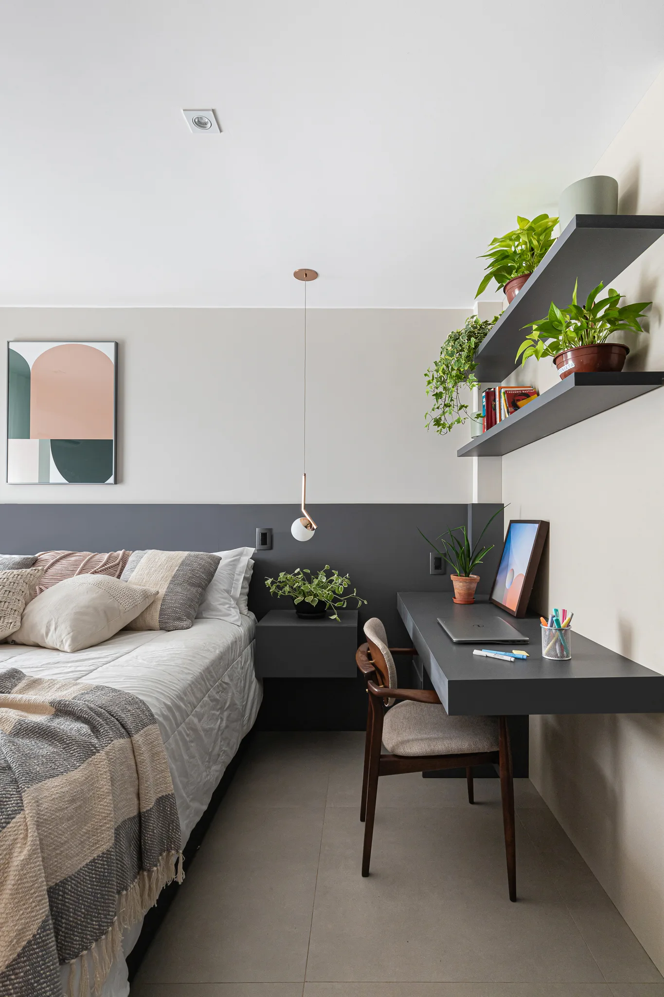 A modern bedroom features a bed with neutral linens, a floating charcoal grey desk with a wood chair, and wall shelves holding potted plants.