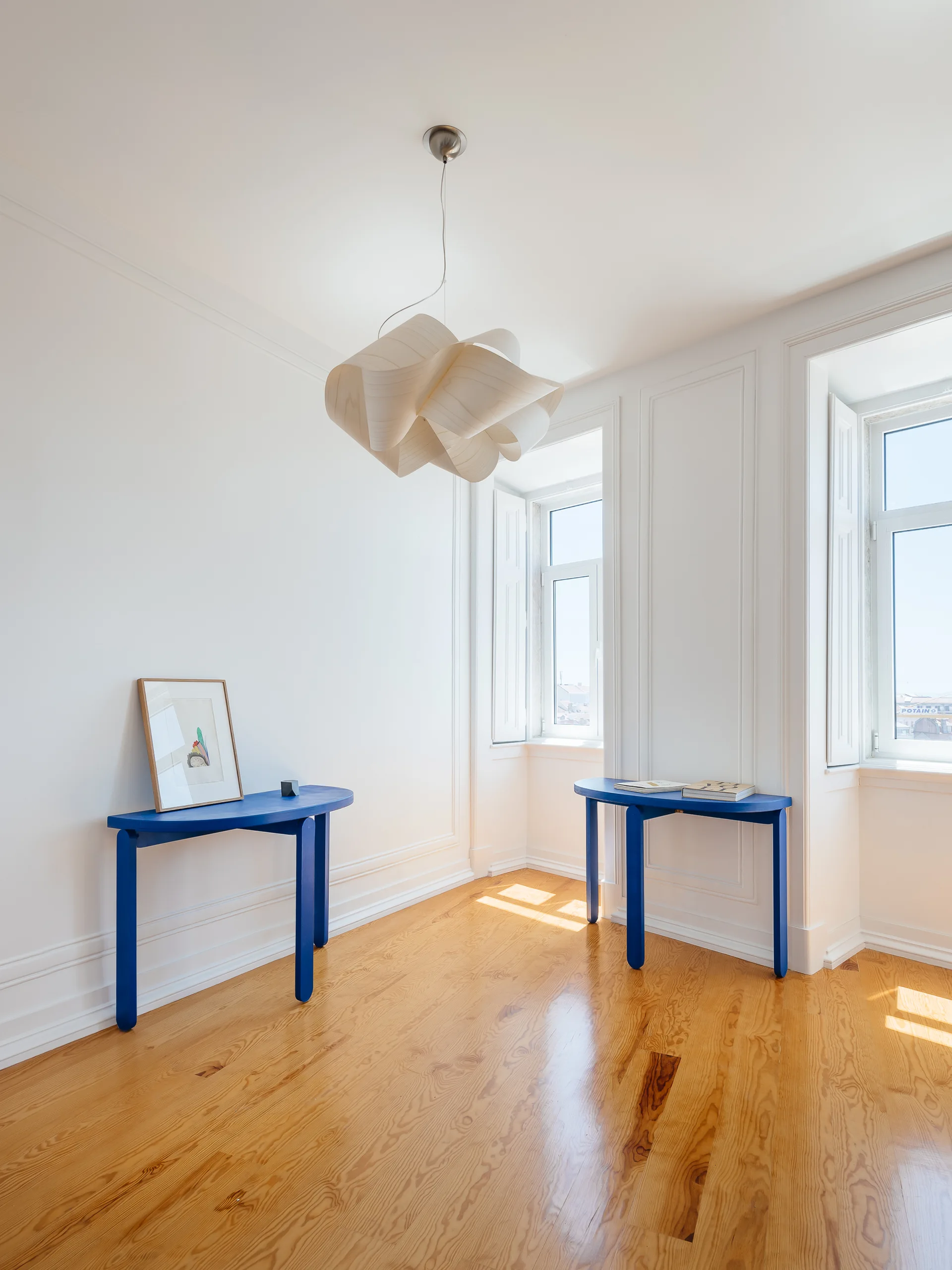 Two cobalt blue console tables sit against white paneled walls in a bright room with hardwood floors and a wavy wood pendant light.