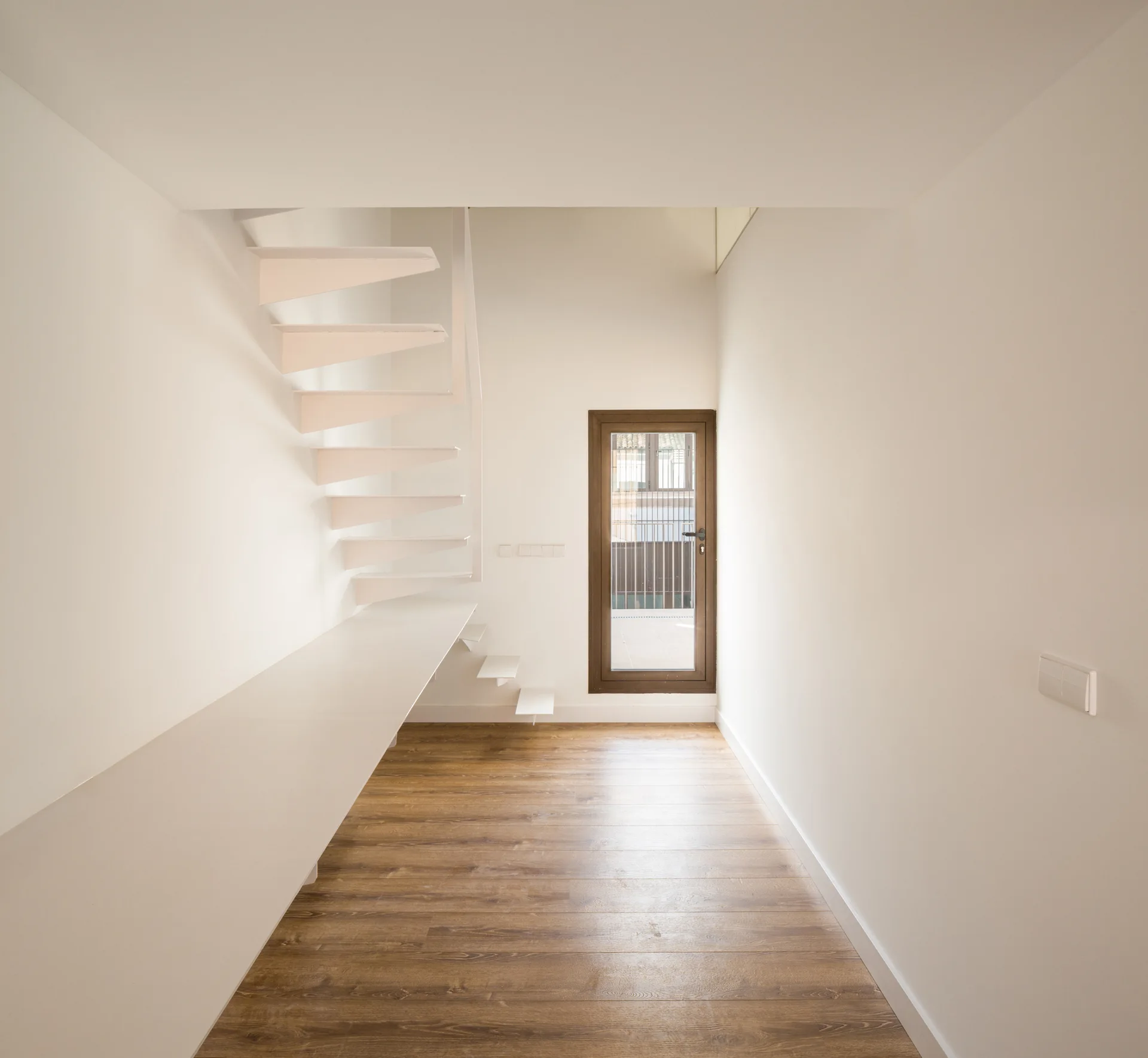 A minimalist white hallway featuring a floating open-tread metal staircase, a long built-in desk, and natural wood flooring.