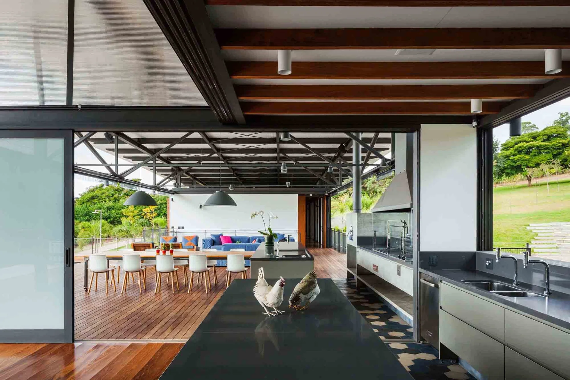 An open-air outdoor kitchen and dining area under a metal truss roof features a long wood table, gray cabinets, and a wood deck.