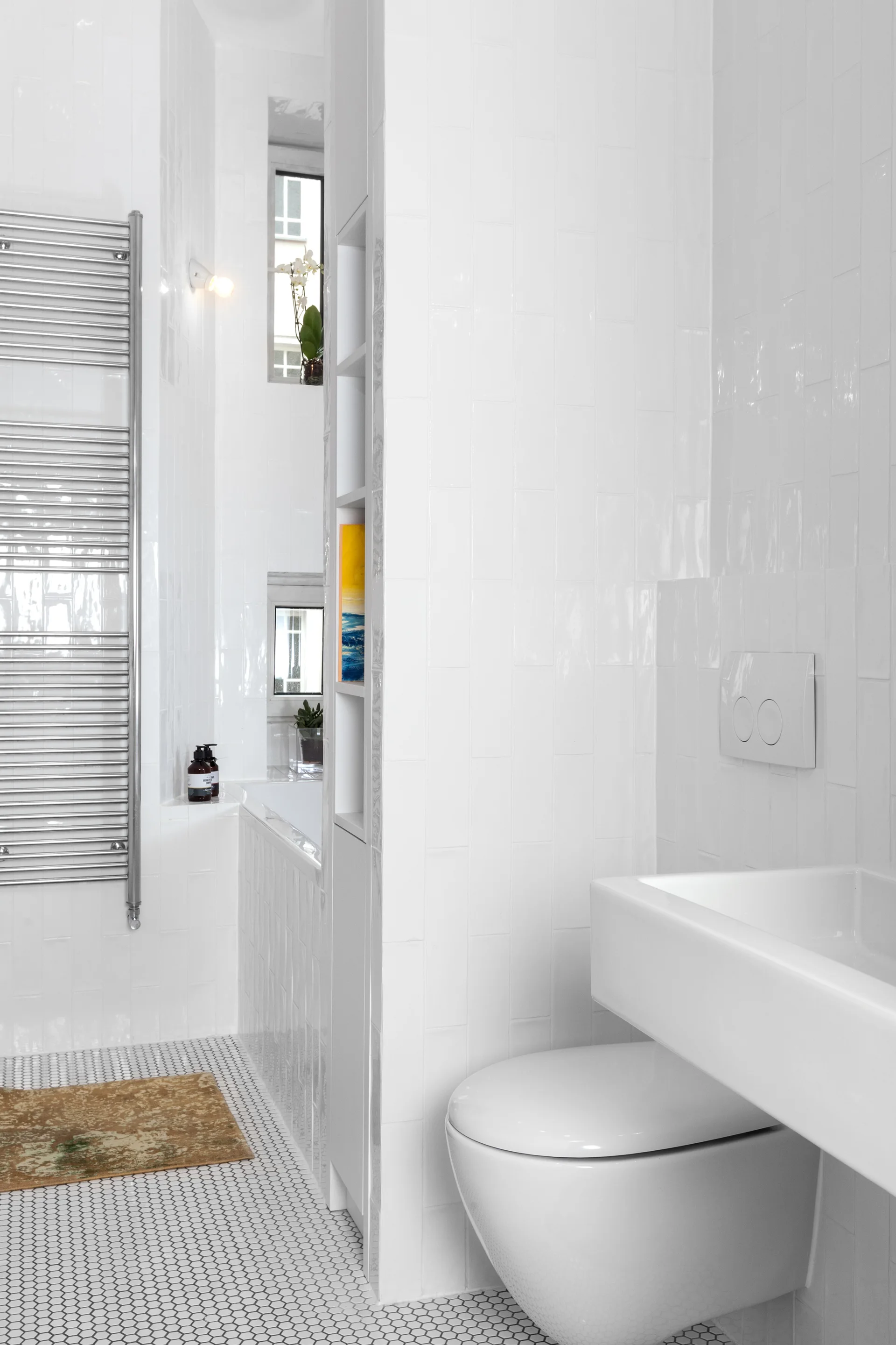 A minimalist white bathroom features vertical subway wall tiles, a small hex tile floor, a wall-mounted toilet, and a narrow built-in shelving unit.