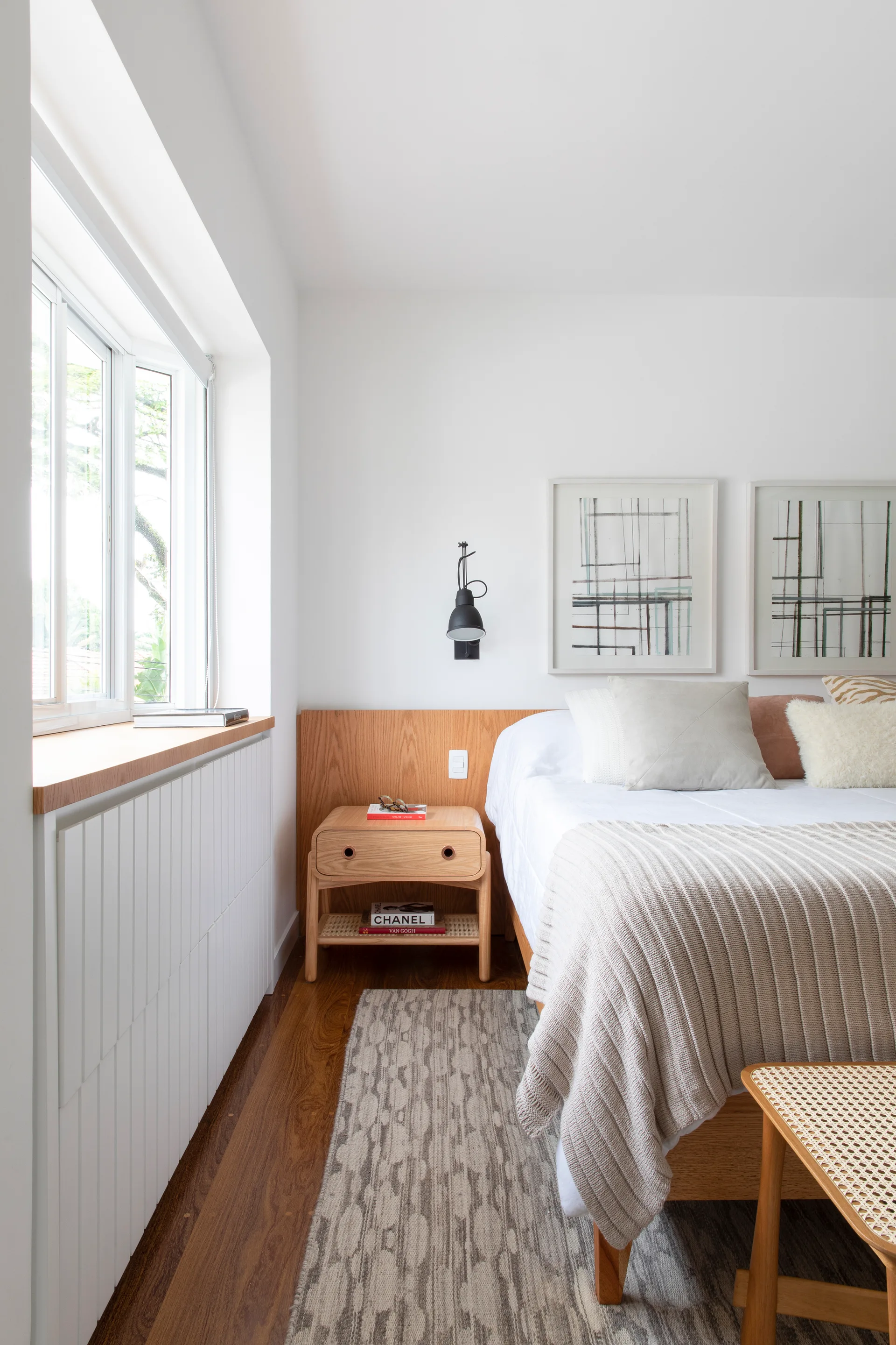 A light-filled bedroom features a low wood headboard panel, a matching oak nightstand, and a white slatted radiator cover beneath a window.