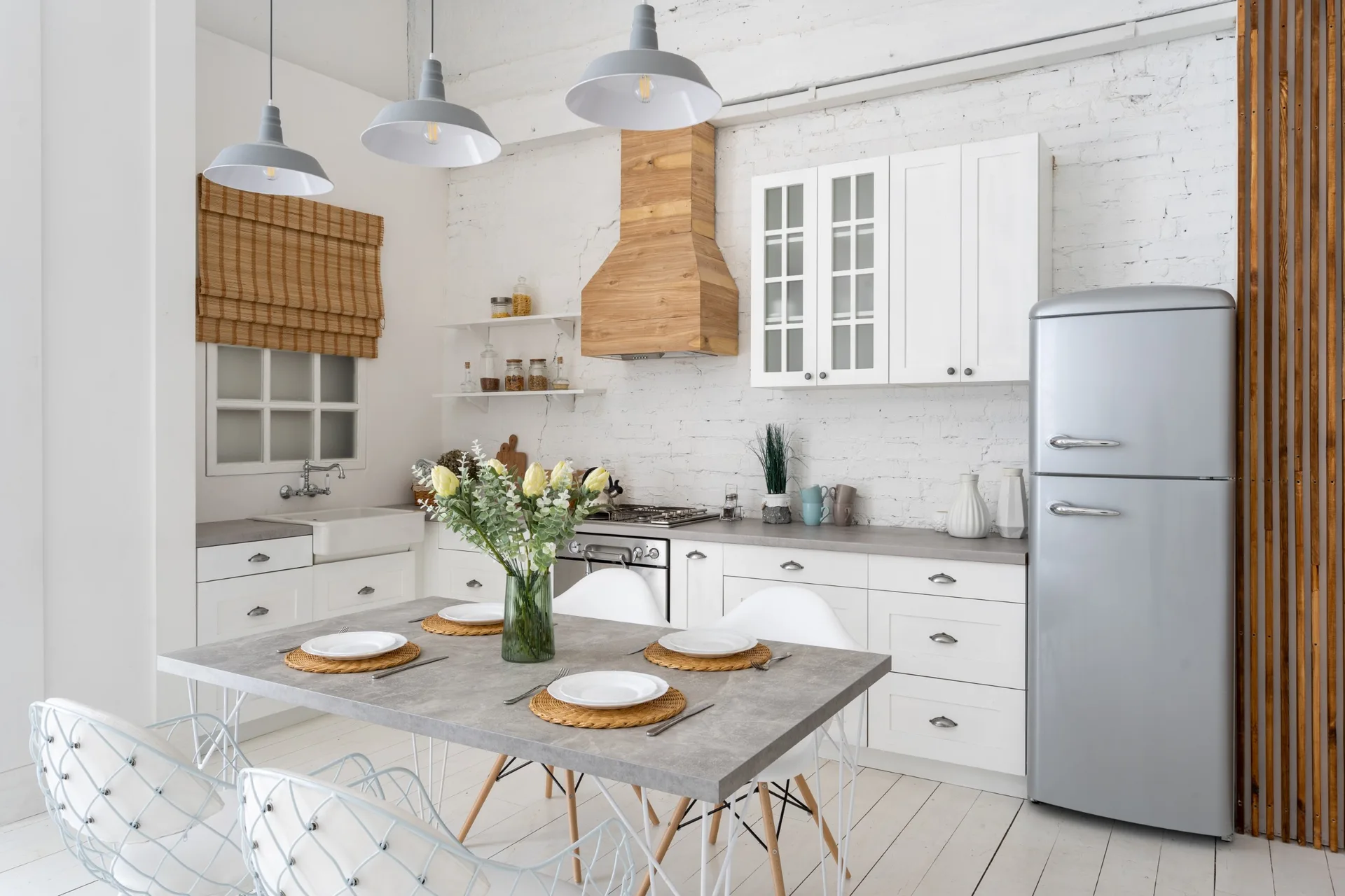 A white kitchen with a gray dining table, white wire chairs, painted brick walls, a natural wood range hood, and a silver retro refrigerator.