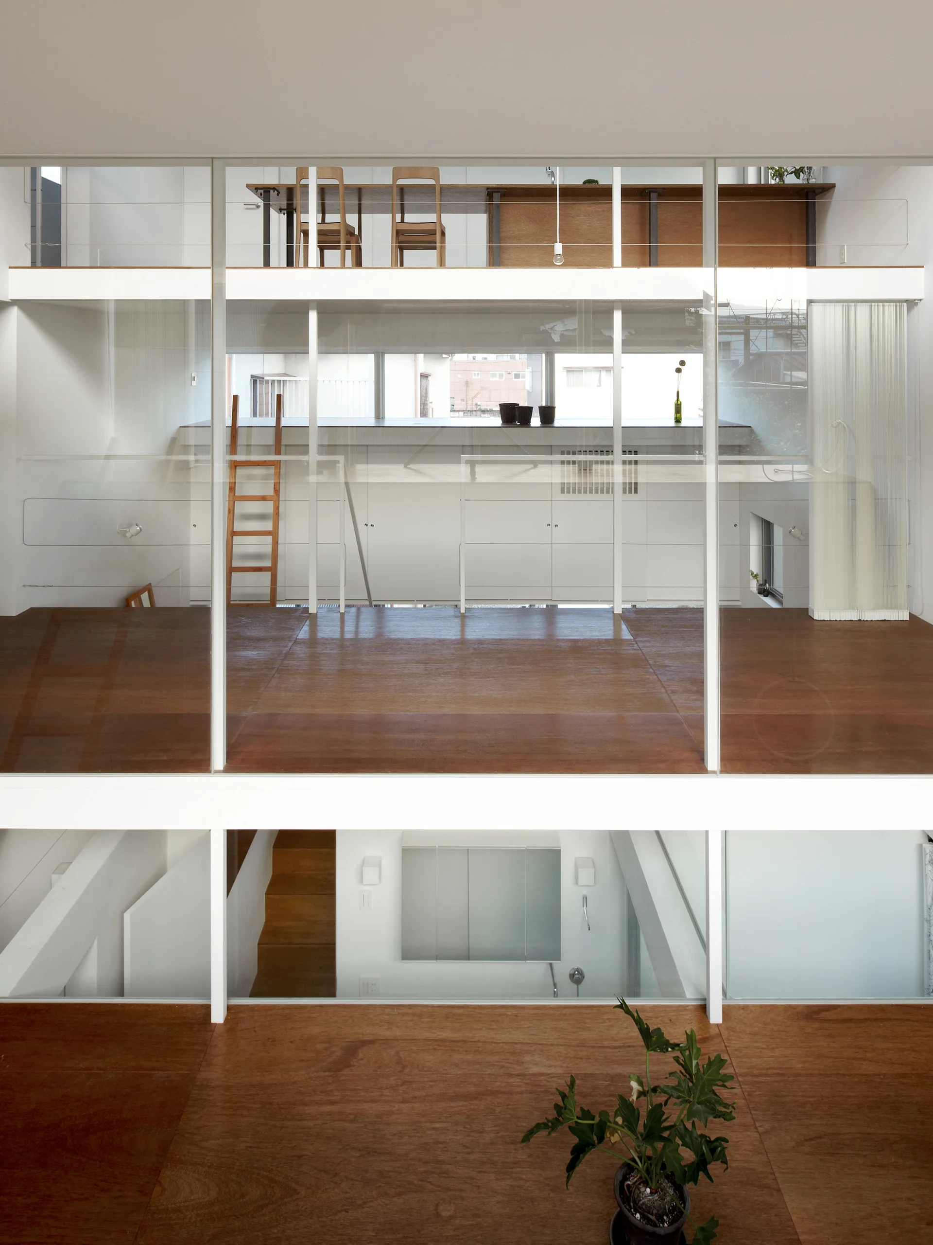 A minimalist multi-level home interior with warm wood floors, white structural framing, transparent glass walls, and built-in storage.