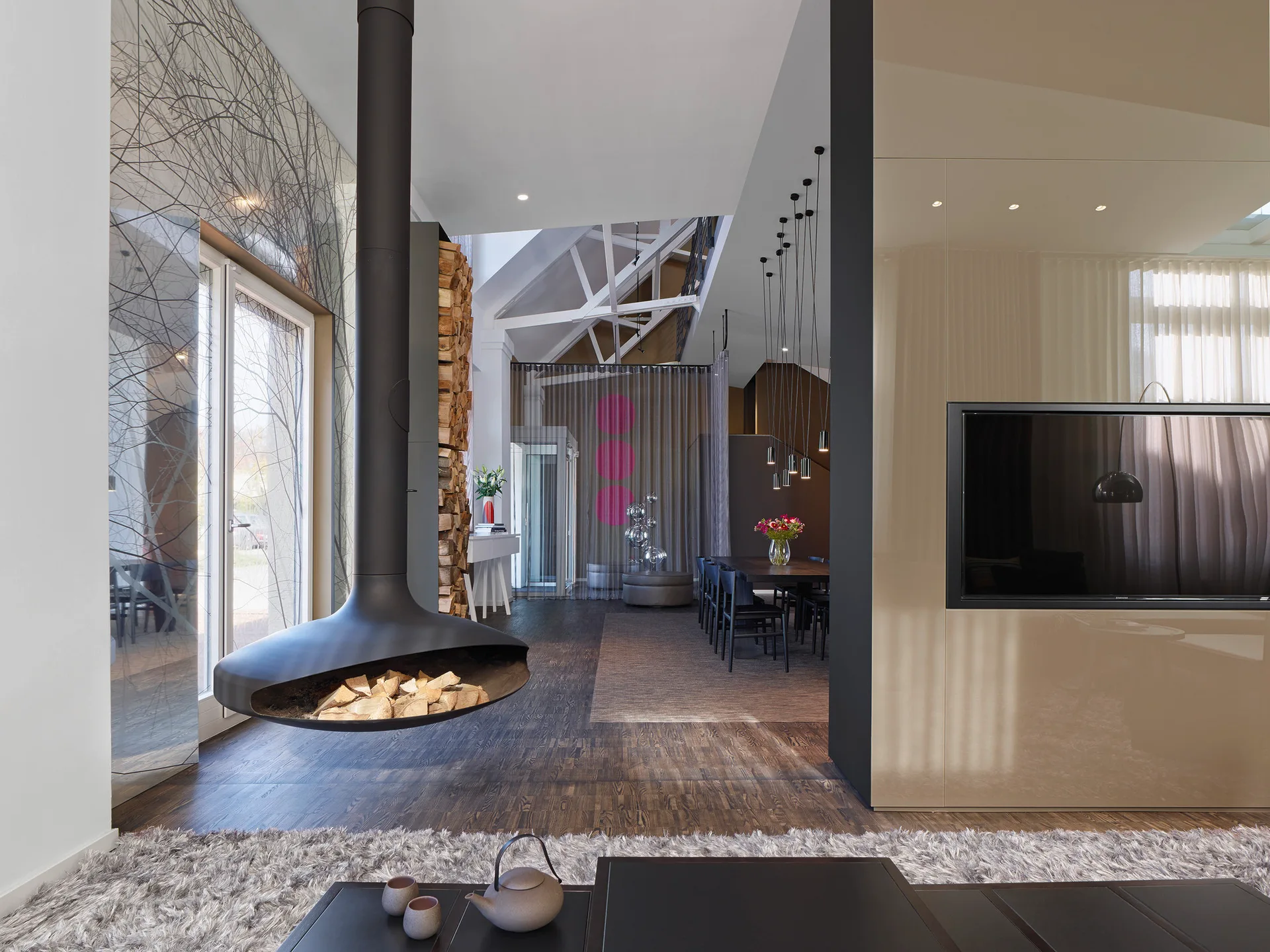 An open-concept living area with a matte black suspended fireplace, high-gloss beige wall panels, a shaggy rug, and a dining table in the background.